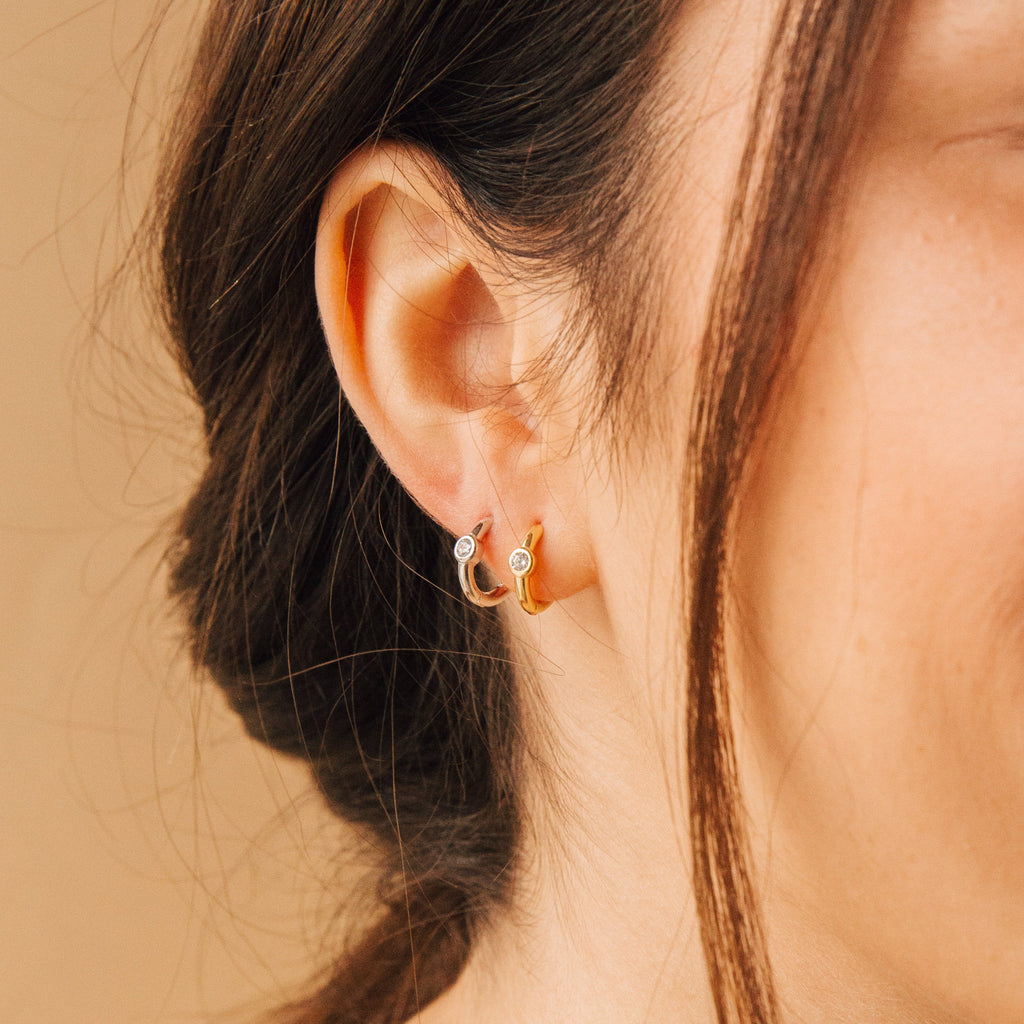 Close-up of a woman's ear wearing Aela Huggies—elegant silver and gold small hoop earrings, each featuring a clear stone accent.
