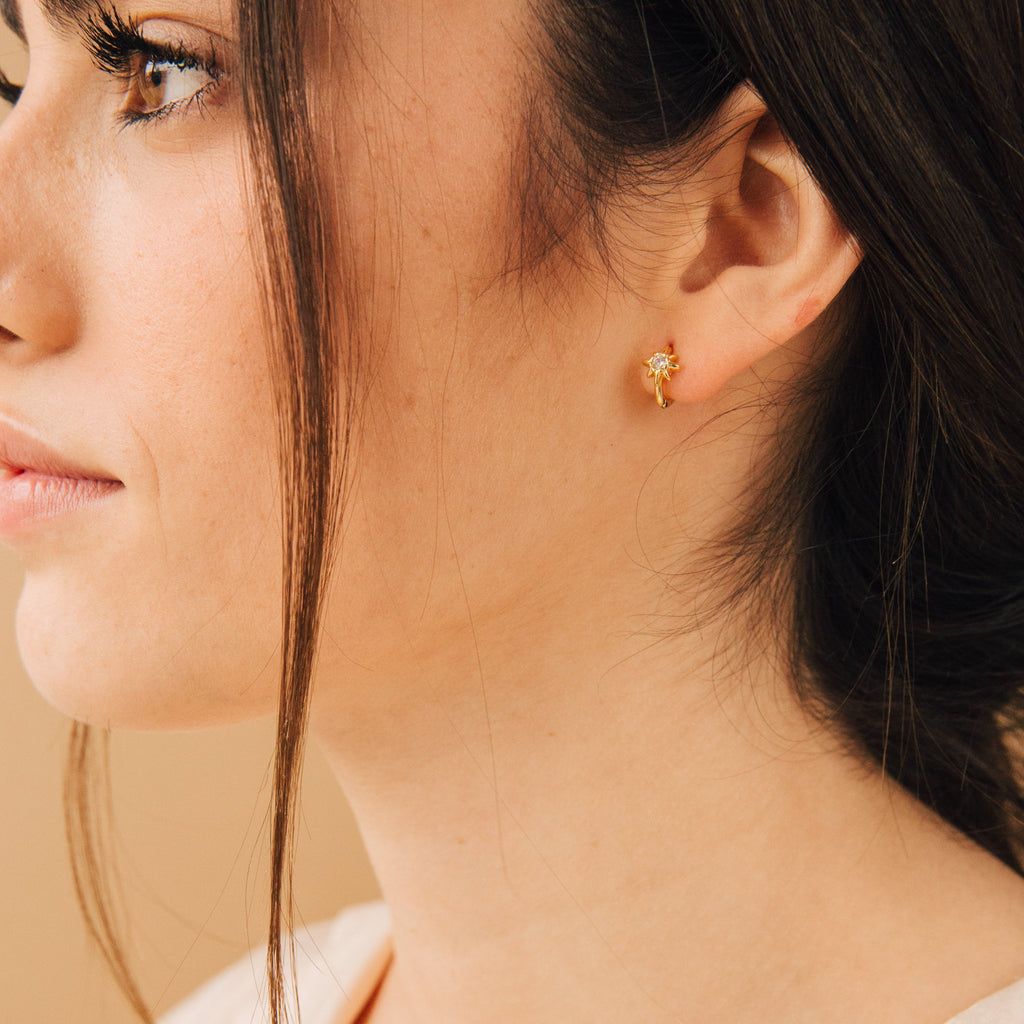 A woman with brown hair is shown in close-up profile wearing the Enya Huggies, small gold flower-shaped earrings. An iridescent diamond catches the light, adding subtle sparkle and elegance to her look.