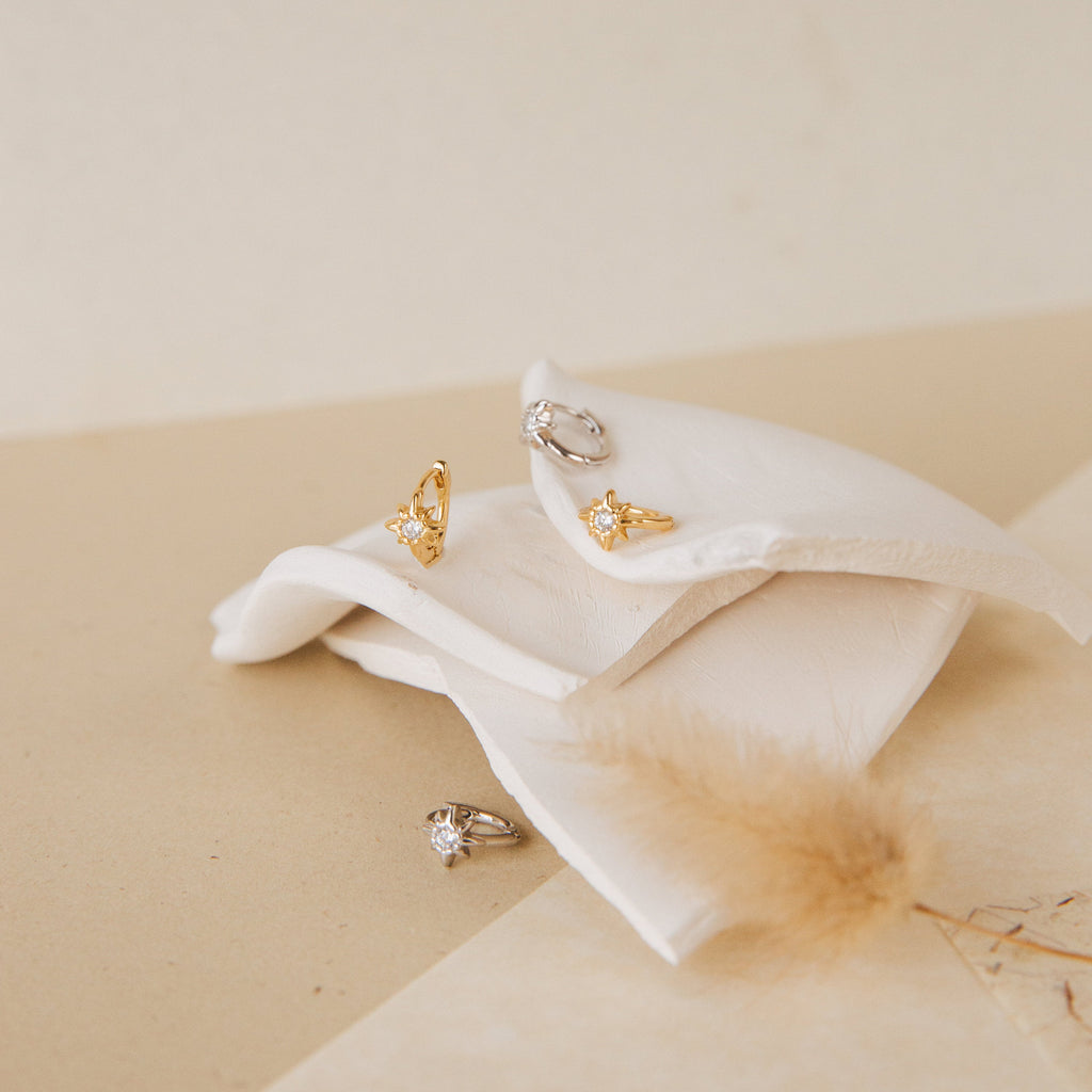The Enya Huggies, gold and silver gemstone rings—one with a star-shaped prong—are displayed on folded white fabric against a soft beige background.