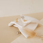 The Enya Huggies, gold and silver gemstone rings—one with a star-shaped prong—are displayed on folded white fabric against a soft beige background.