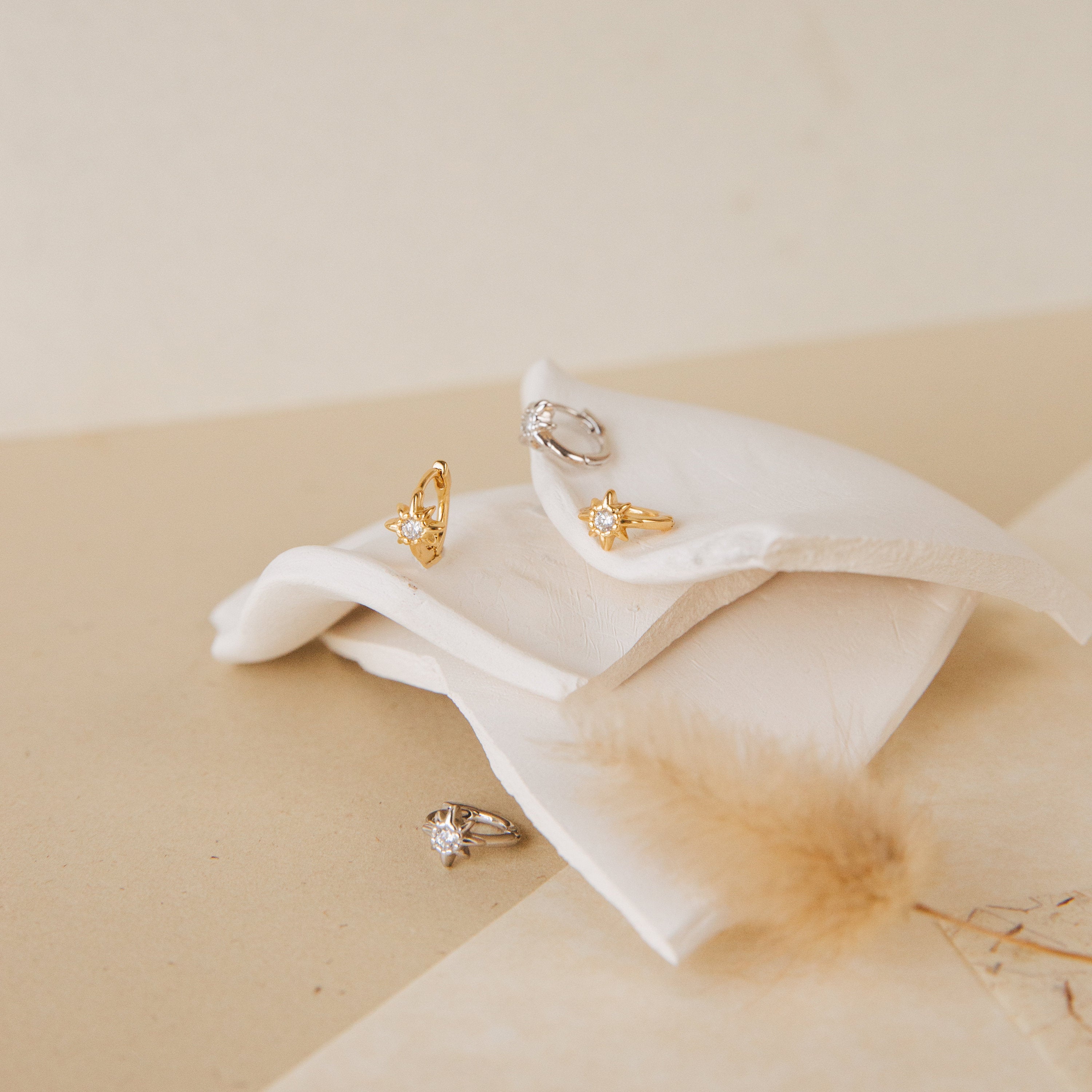 The Enya Huggies, gold and silver gemstone rings—one with a star-shaped prong—are displayed on folded white fabric against a soft beige background.