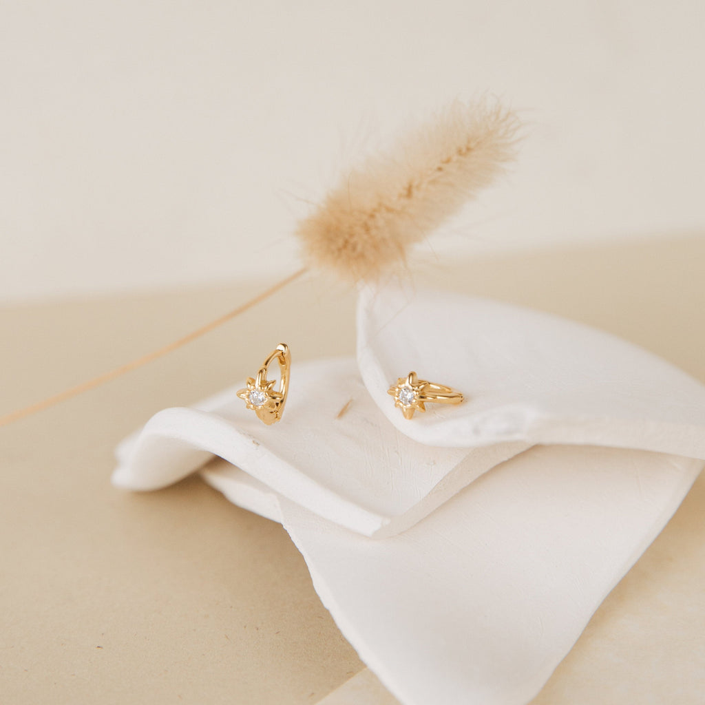Enya Huggies gold earrings with star-shaped prong designs rest on folded white fabric, accented by a dried grass stem—ideal for those who love delicate details and iridescent diamond sparkle.