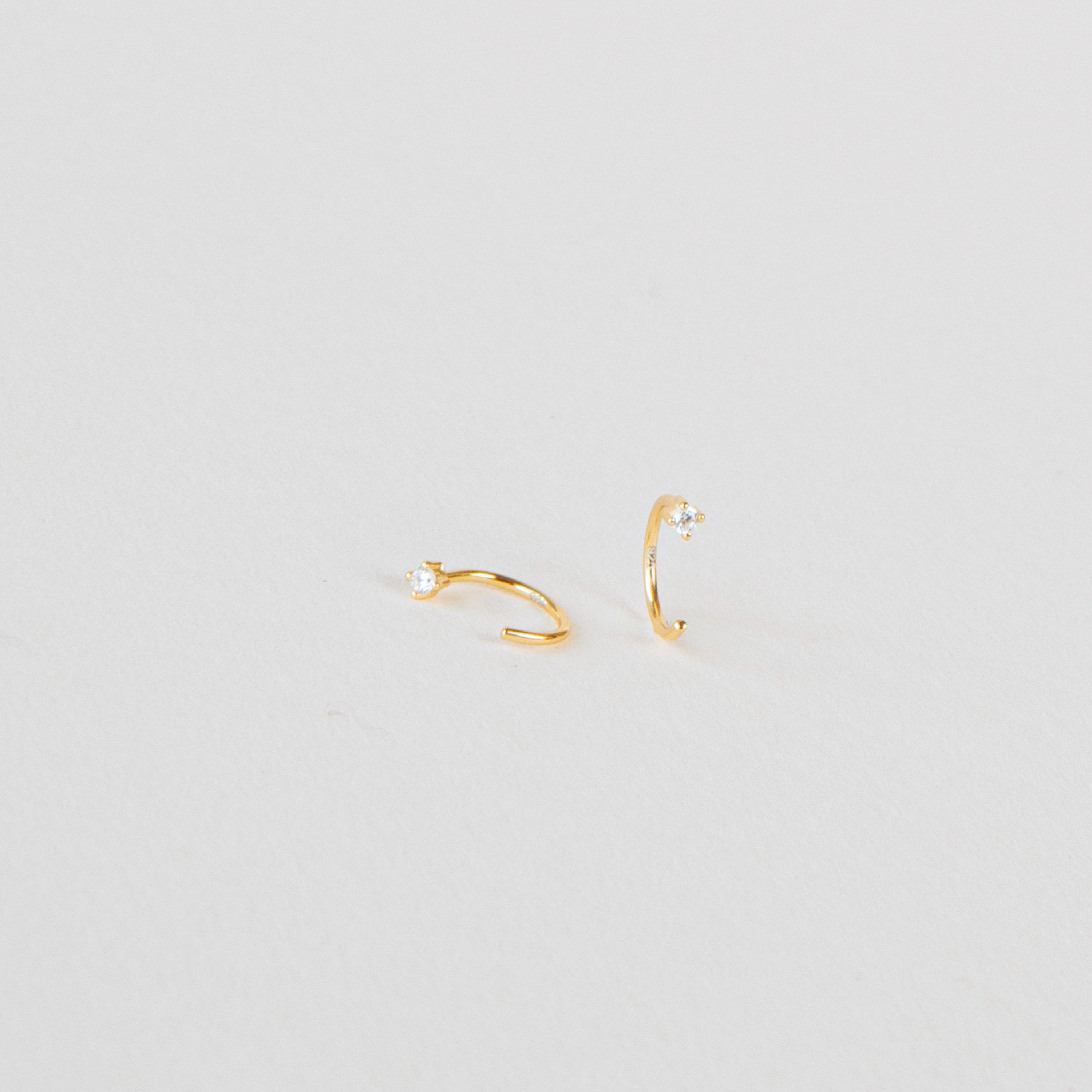 The Saylor Huggies are two small gold hoop nose rings adorned with tiny clear gemstones, displayed on a plain white background.