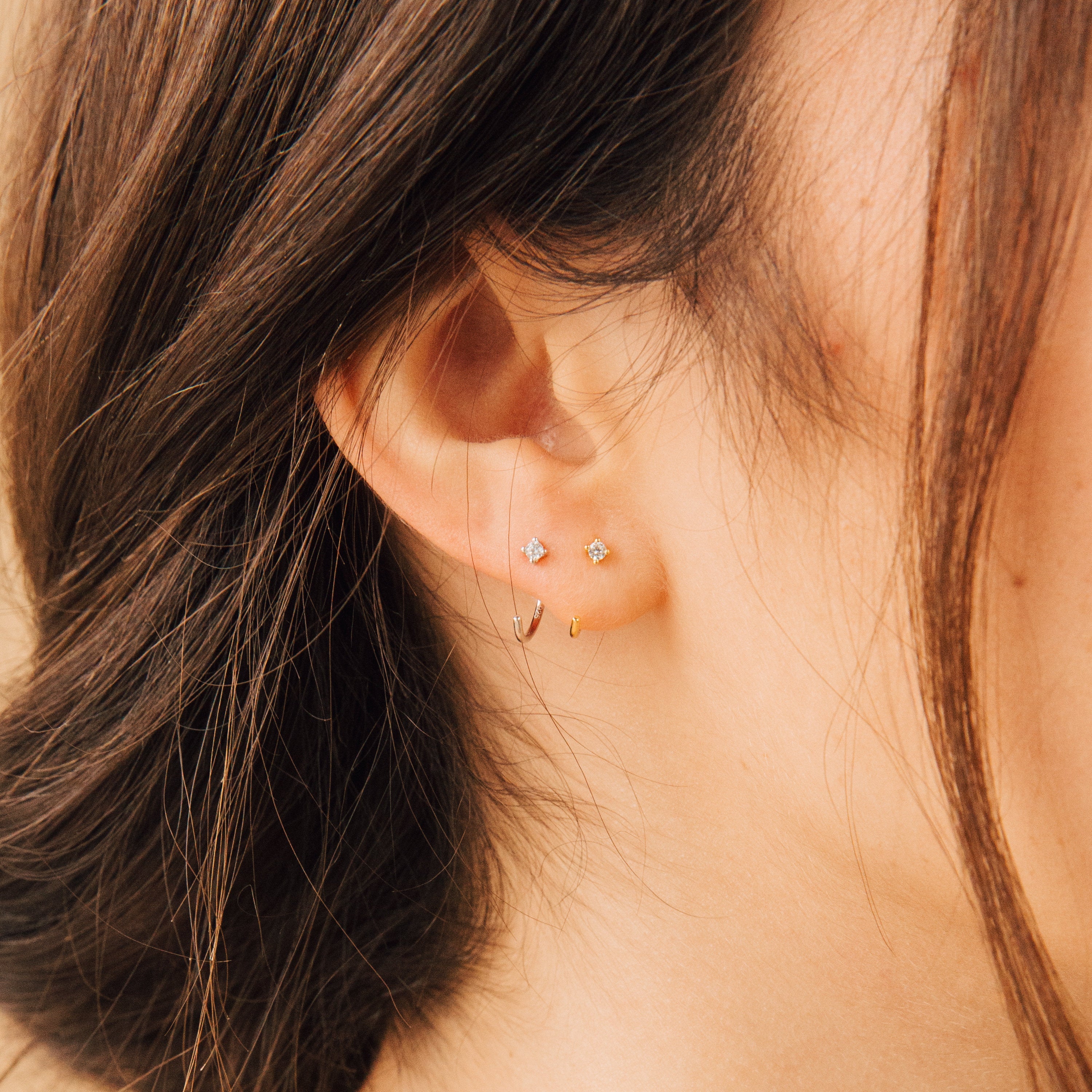 Close-up of a woman's ear adorned with two small studs and the Saylor Huggies, showcasing a graceful mix of delicate earrings.
