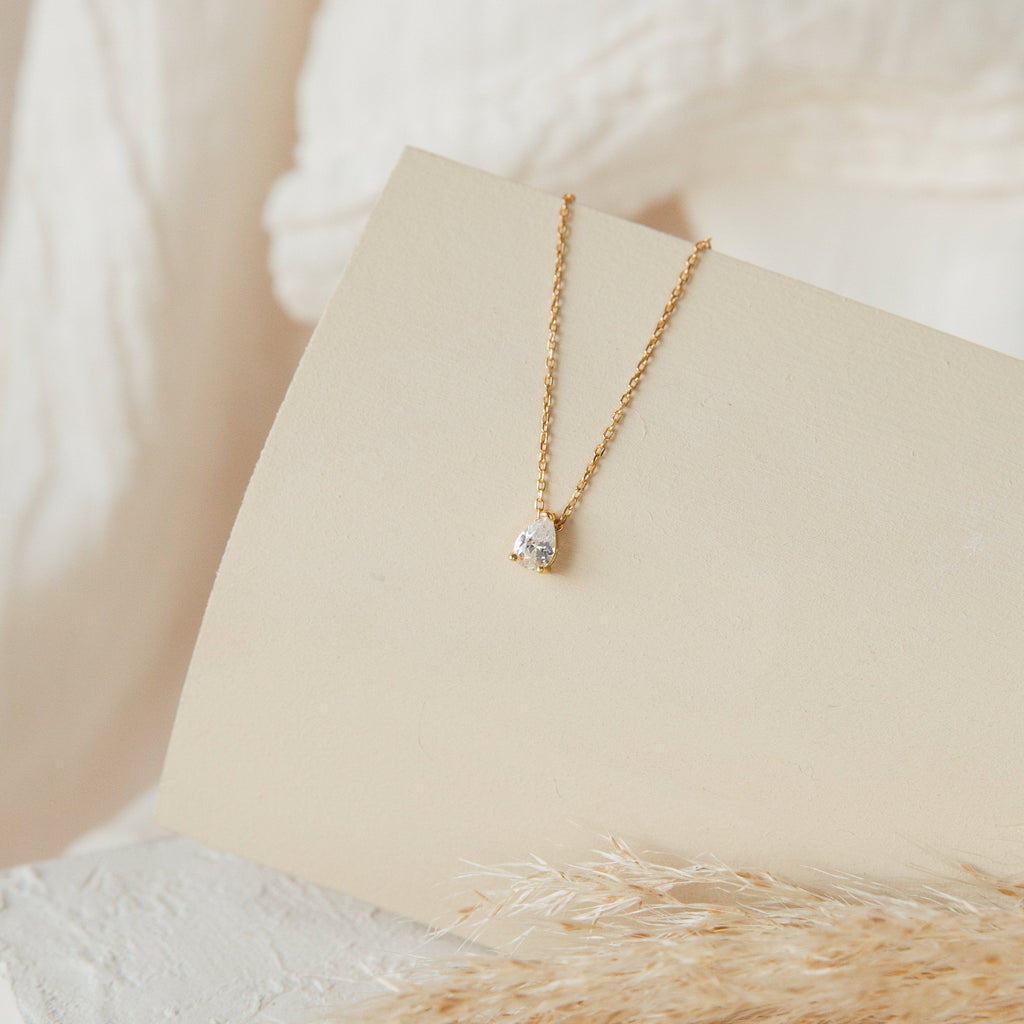 Rae Diamond Necklace with solitaire diamond pendant on 18K gold chain on cream background.