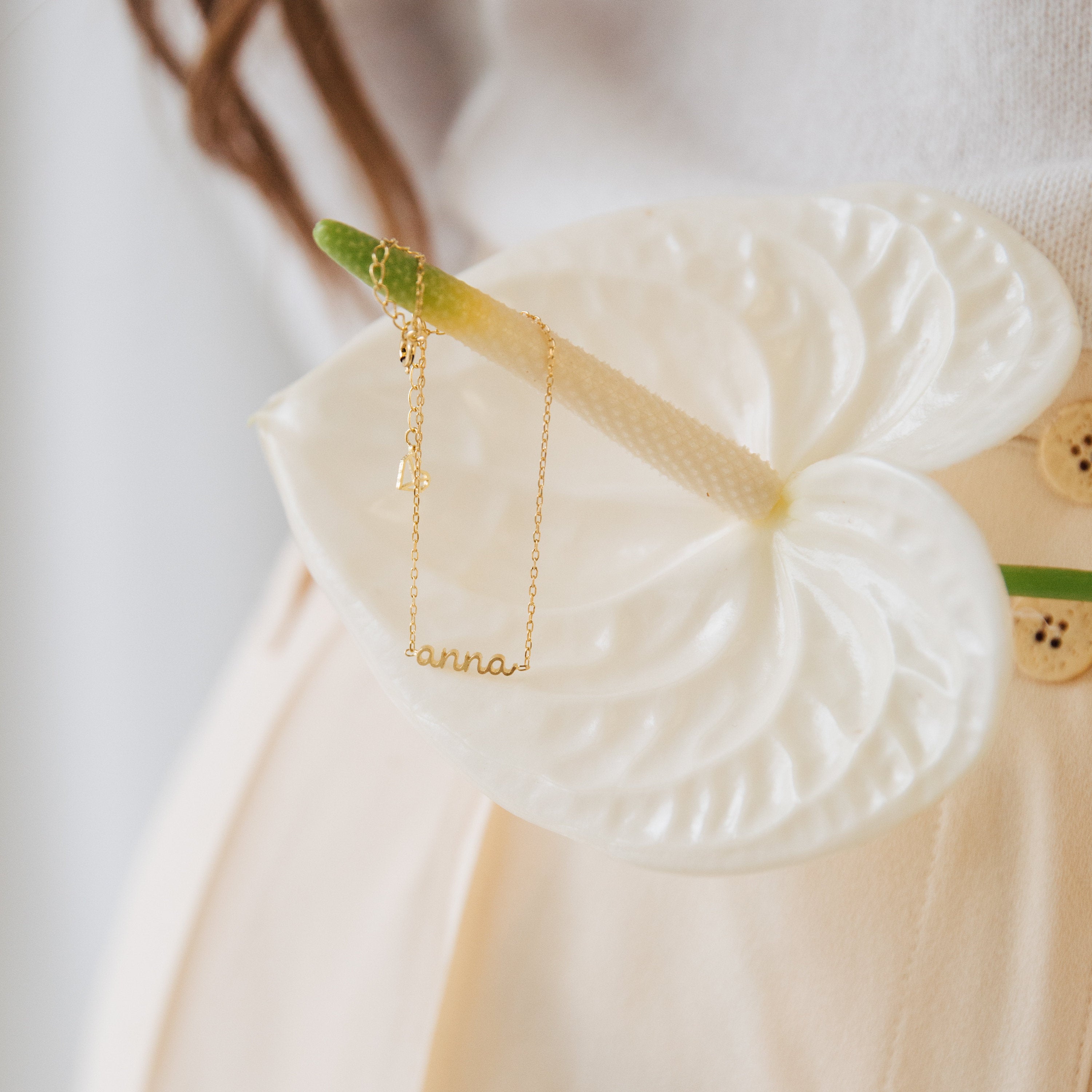 The Petite Tiny Name Necklace with "anna" is shown hanging from a white anthurium flower on a light background—a delicate, personalized gift that's perfect for mom.