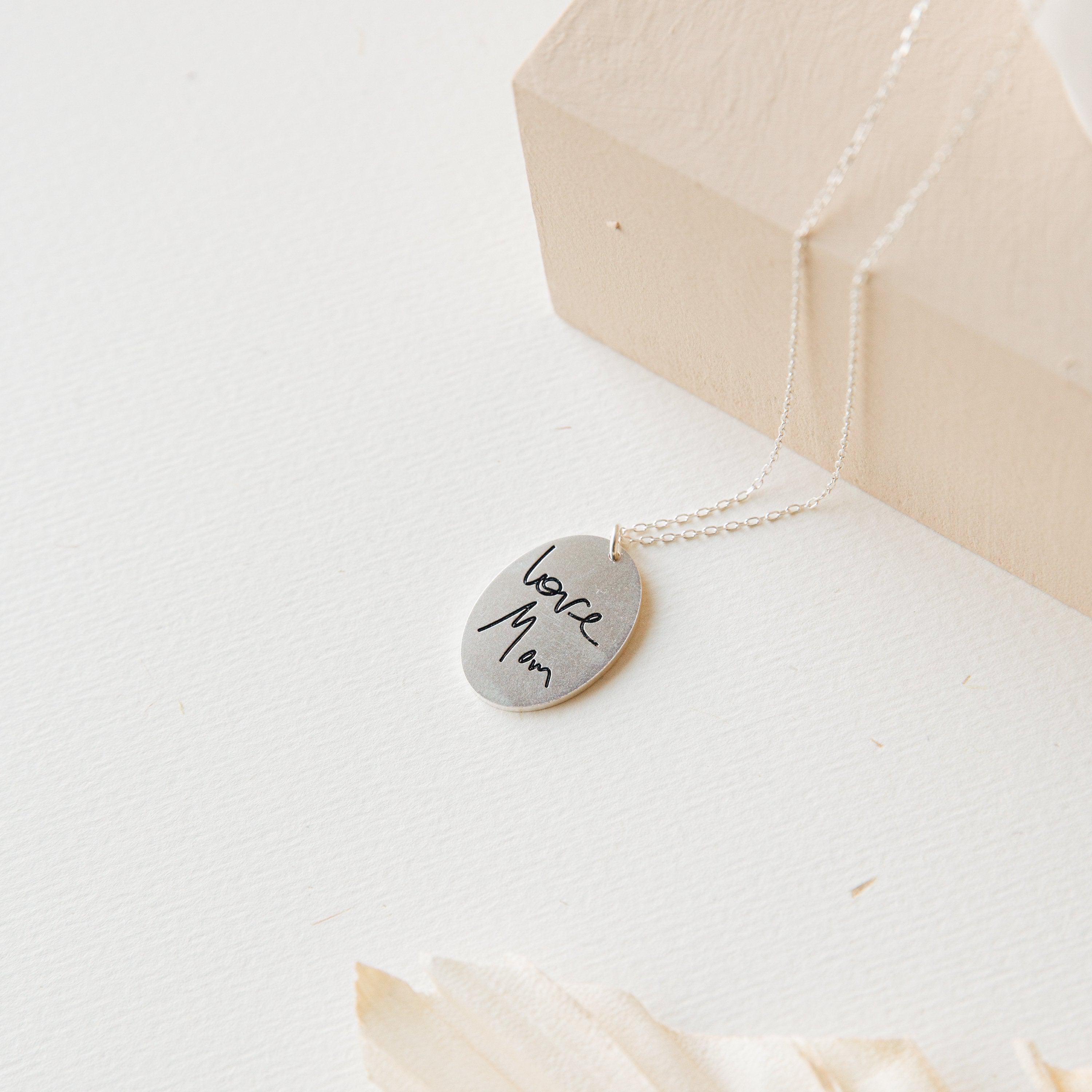 The Oval Fingerprint Necklace is a silver oval pendant, engraved with "love Mom" and personalized fingerprint etching—a heartfelt memorial jewelry keepsake shown on a light surface with a soft background.