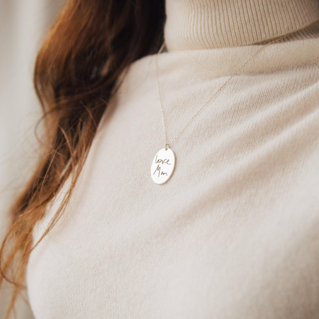 A woman in a beige turtleneck wears an Oval Fingerprint Necklace featuring personalized etching that reads "love Mom," highlighting elegant memorial jewelry.
