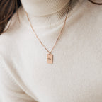 Close-up of the Name Tag Necklace in rose gold, featuring a rectangular "Muse" pendant, worn over a cream turtleneck sweater.