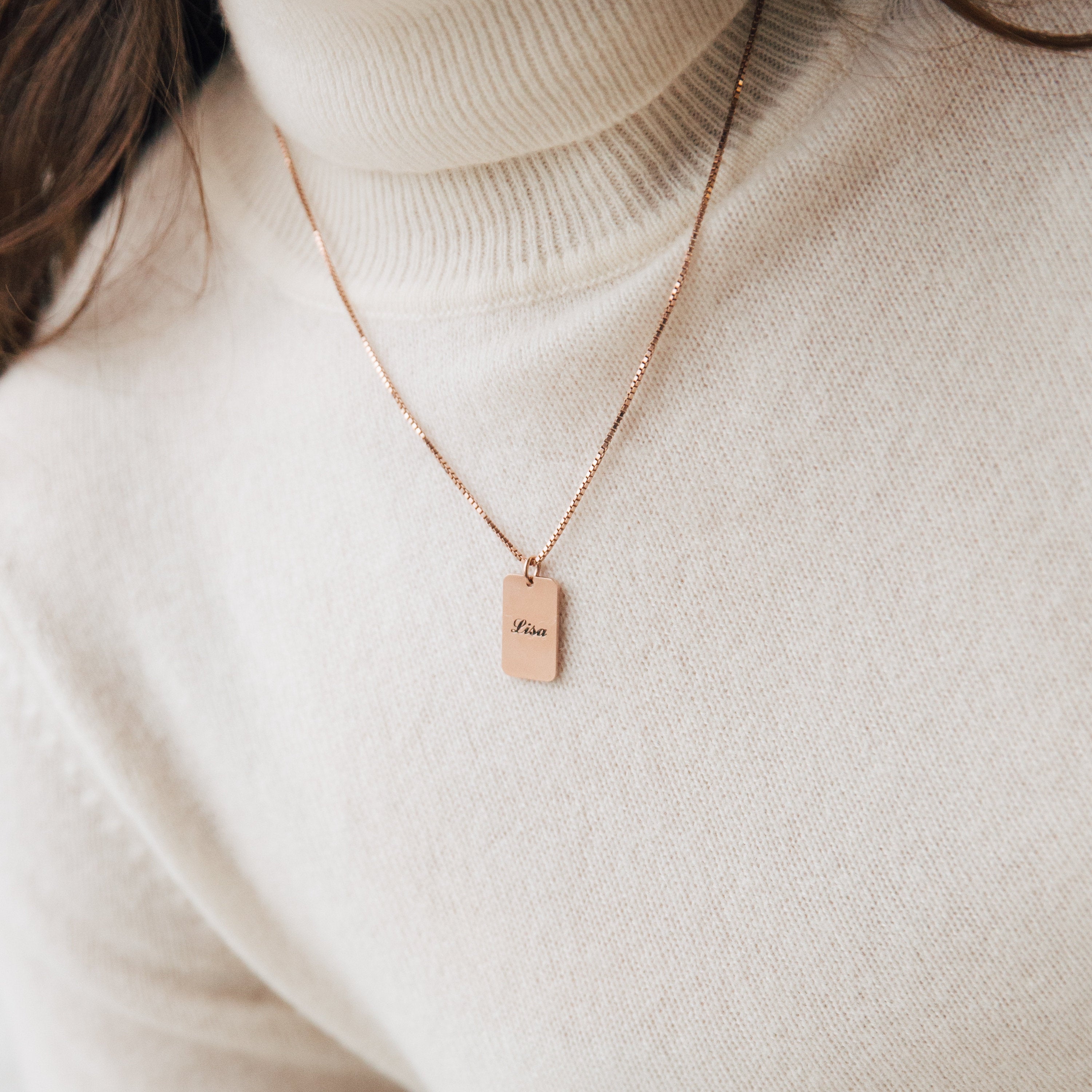 Close-up of the Name Tag Necklace in rose gold, featuring a rectangular "Muse" pendant, worn over a cream turtleneck sweater.