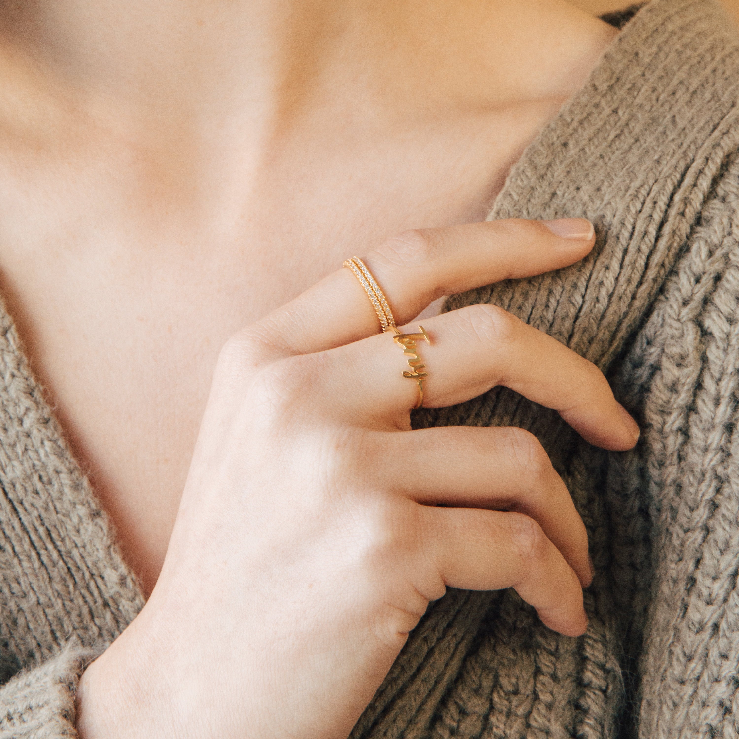 A hand wearing the Grace Script Name Ring in Rose Gold and another gold band gently touches a beige knitted sweater near the collarbone.