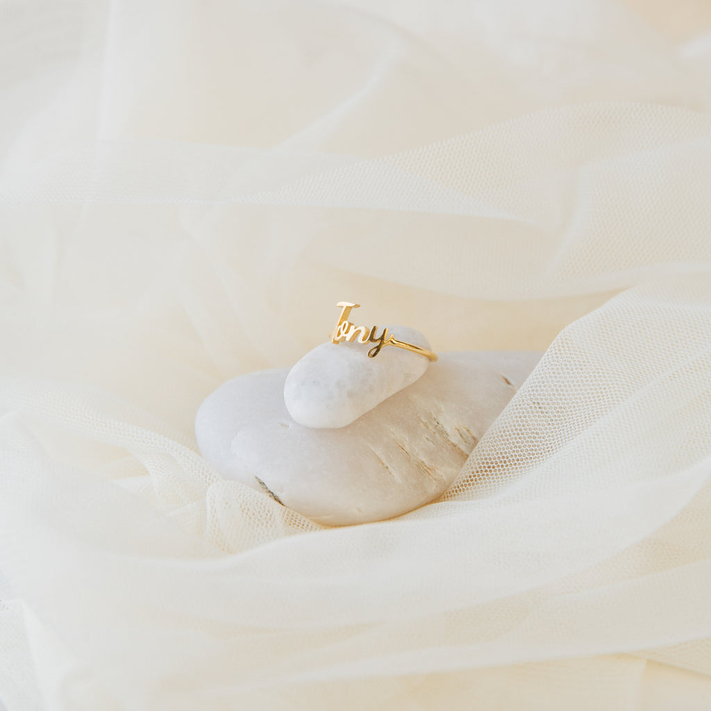 The Grace Script Name Ring in Rose Gold, personalized with the name Tony, is displayed on a white stone and surrounded by soft beige fabric.