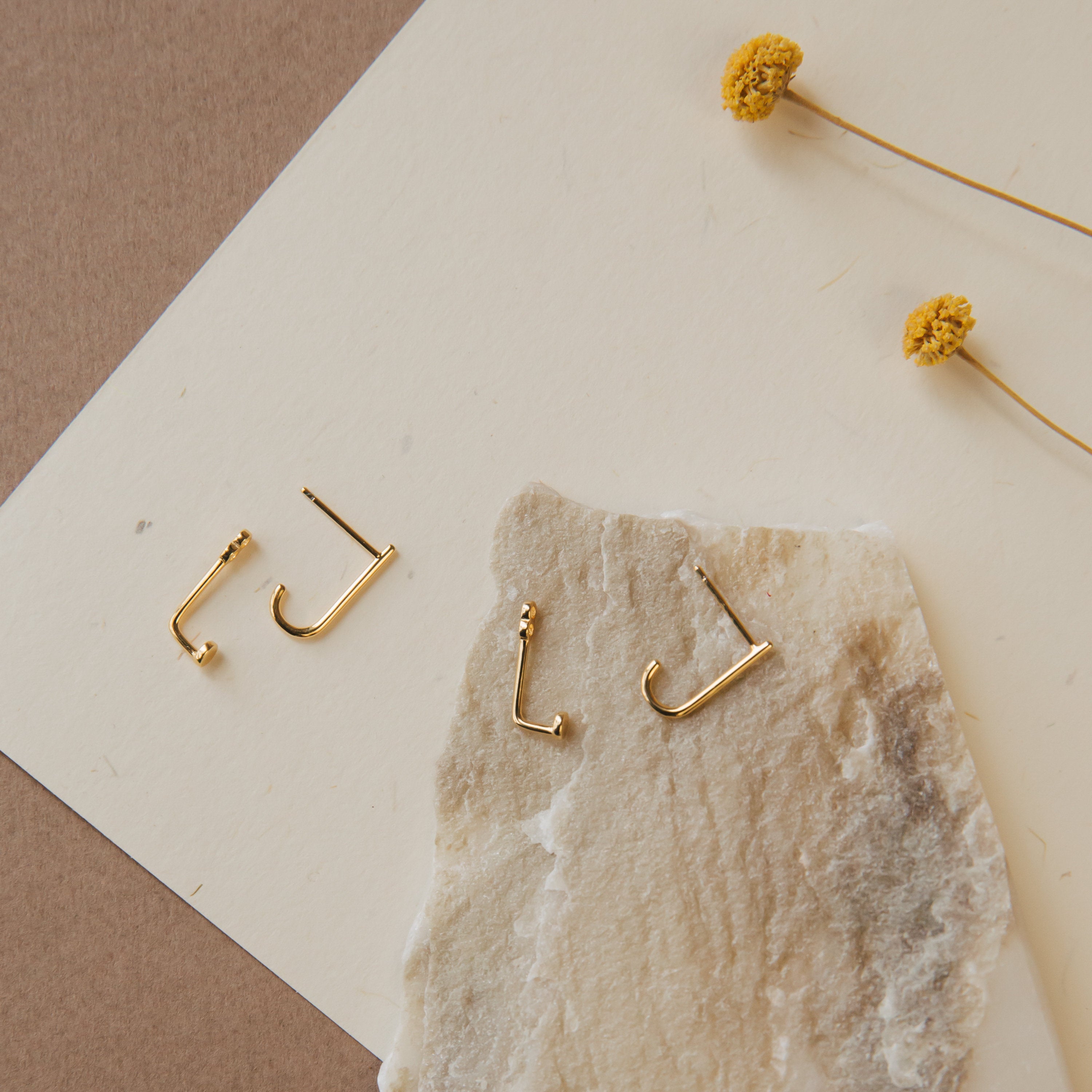 Two pairs of Dot Ear Jackets are displayed on textured paper with two yellow dried flowers nearby, highlighting the modern earrings' minimalist geometric style.