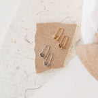 Two pairs of minimalist Ava Earrings—one gold, one silver, including chic safety pin designs—are displayed on textured brown paper.
