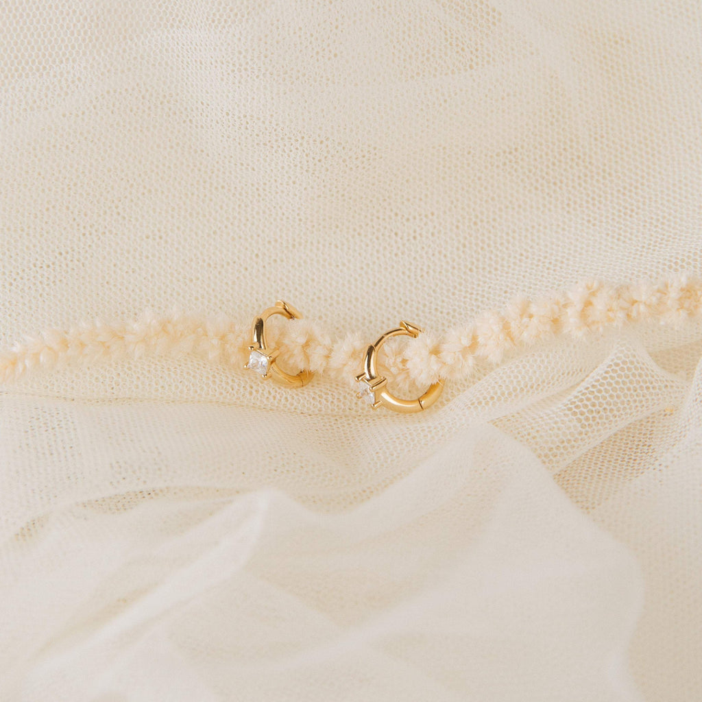 The Athena Huggies, a small gold hoop earring with a tiny gemstone, rests on soft cream-colored fabric.