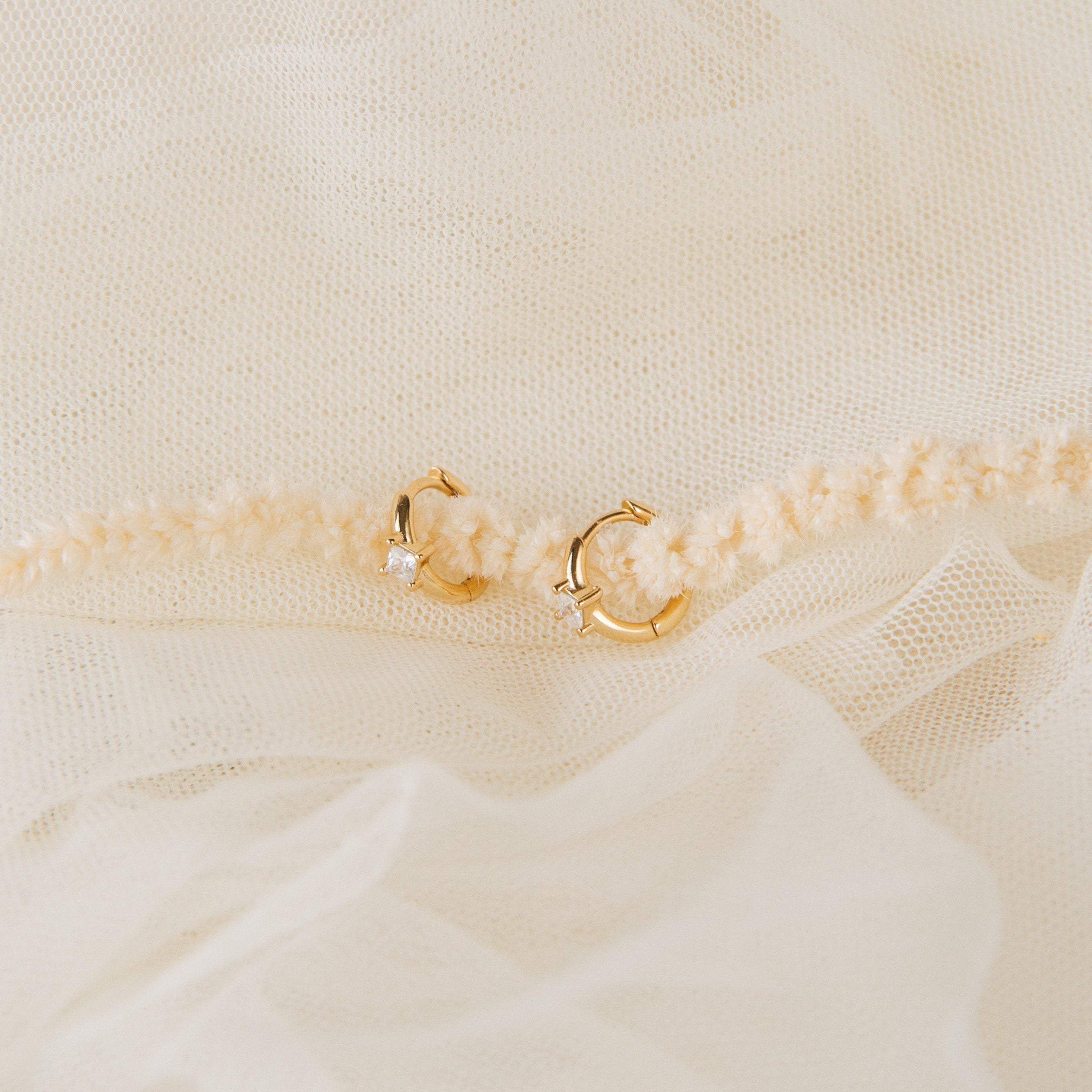 The Athena Huggies, a small gold hoop earring with a tiny gemstone, rests on soft cream-colored fabric.