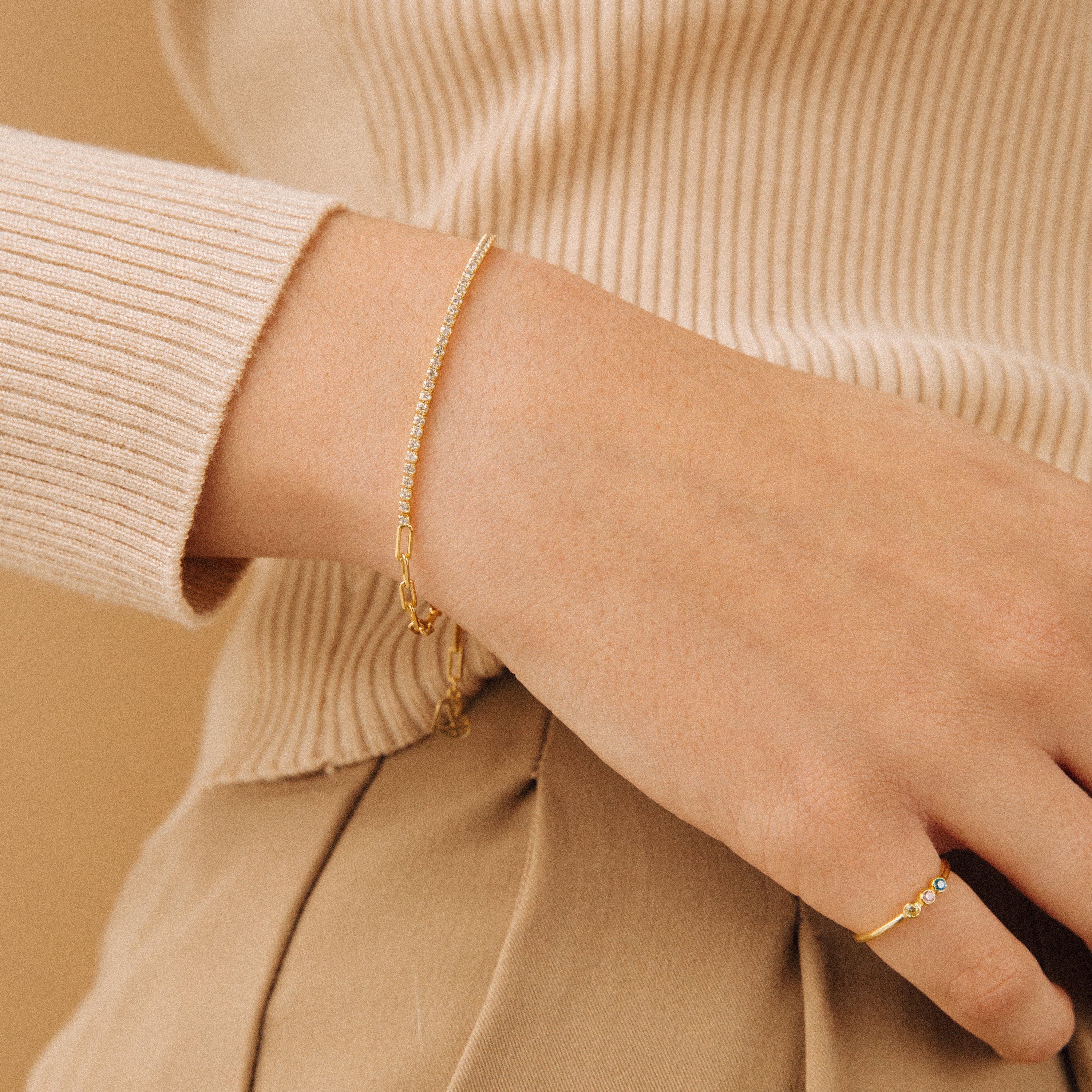 A hand wearing the Klara Tennis Bracelet and a gold ring with small gems is styled against a beige outfit for a chic minimalist jewelry look.