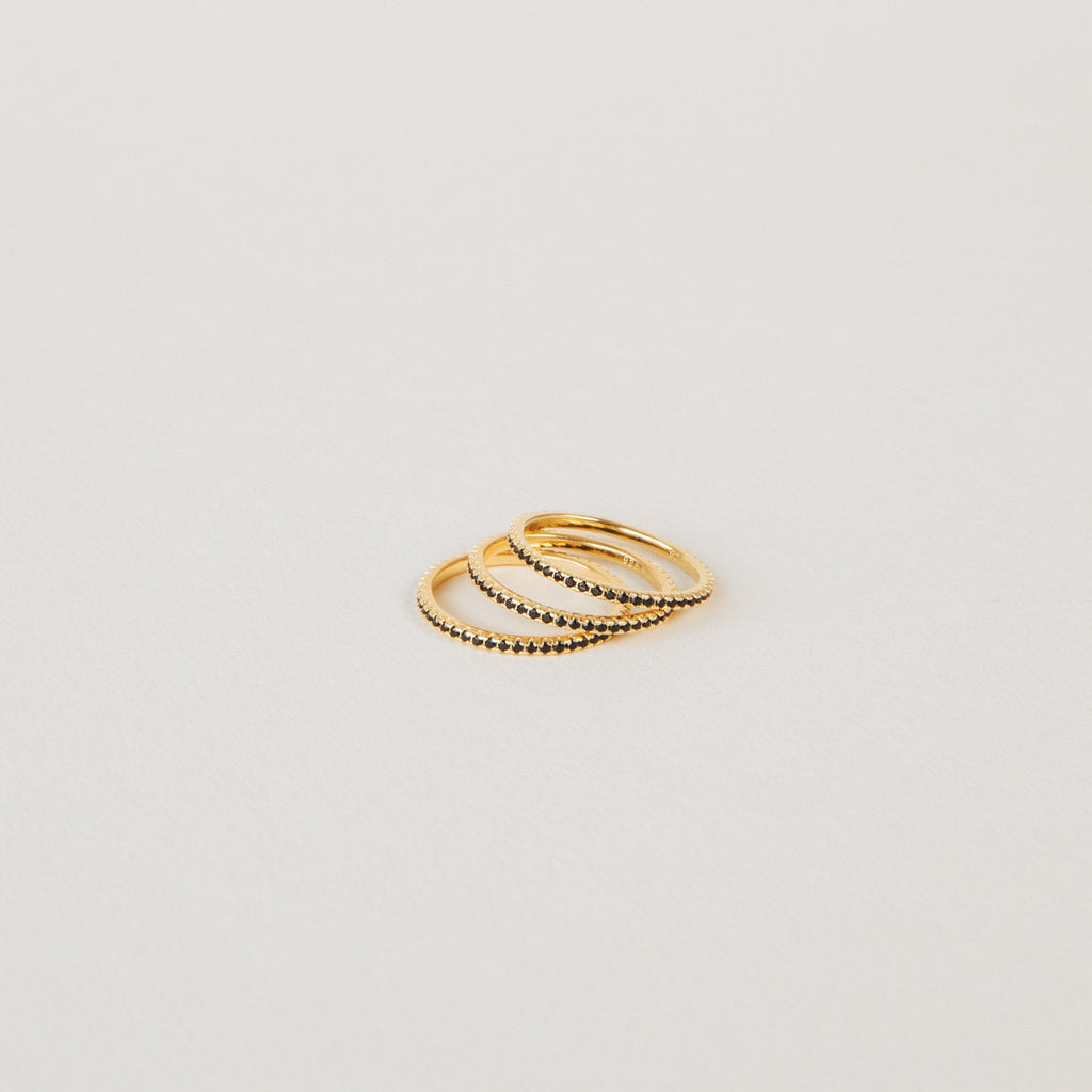 The Amber Eternity Ring, a thin gold beaded stackable ring, is arranged on a plain light-colored surface with two other slim gold rings.