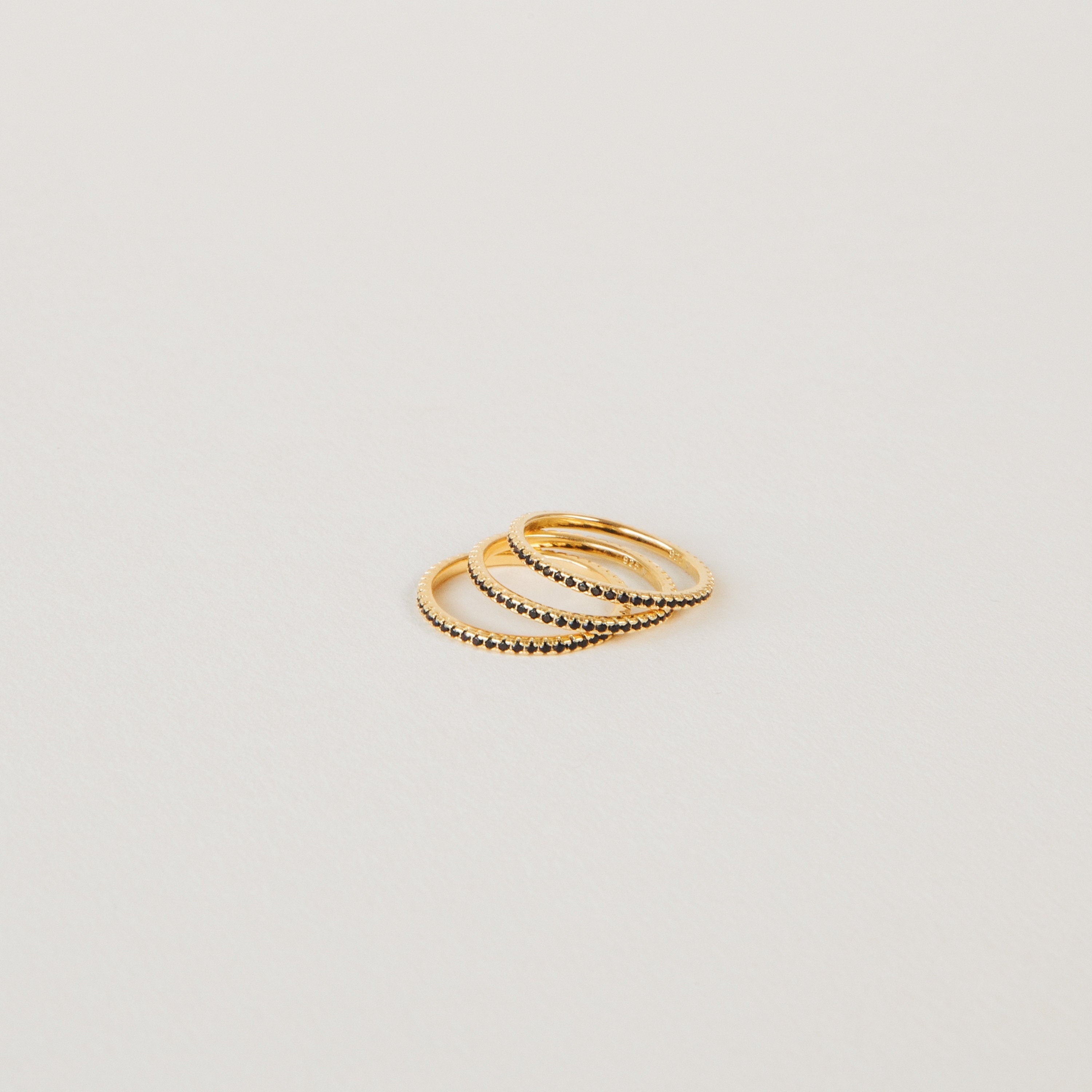 The Amber Eternity Ring, a thin gold beaded stackable ring, is arranged on a plain light-colored surface with two other slim gold rings.