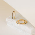 The Amber Eternity Ring, a delicate gold stackable ring with beaded edges, is displayed on a white stone surface against a neutral background.