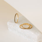 The Amber Eternity Ring, a delicate gold stackable ring with beaded edges, is displayed on a white stone surface against a neutral background.