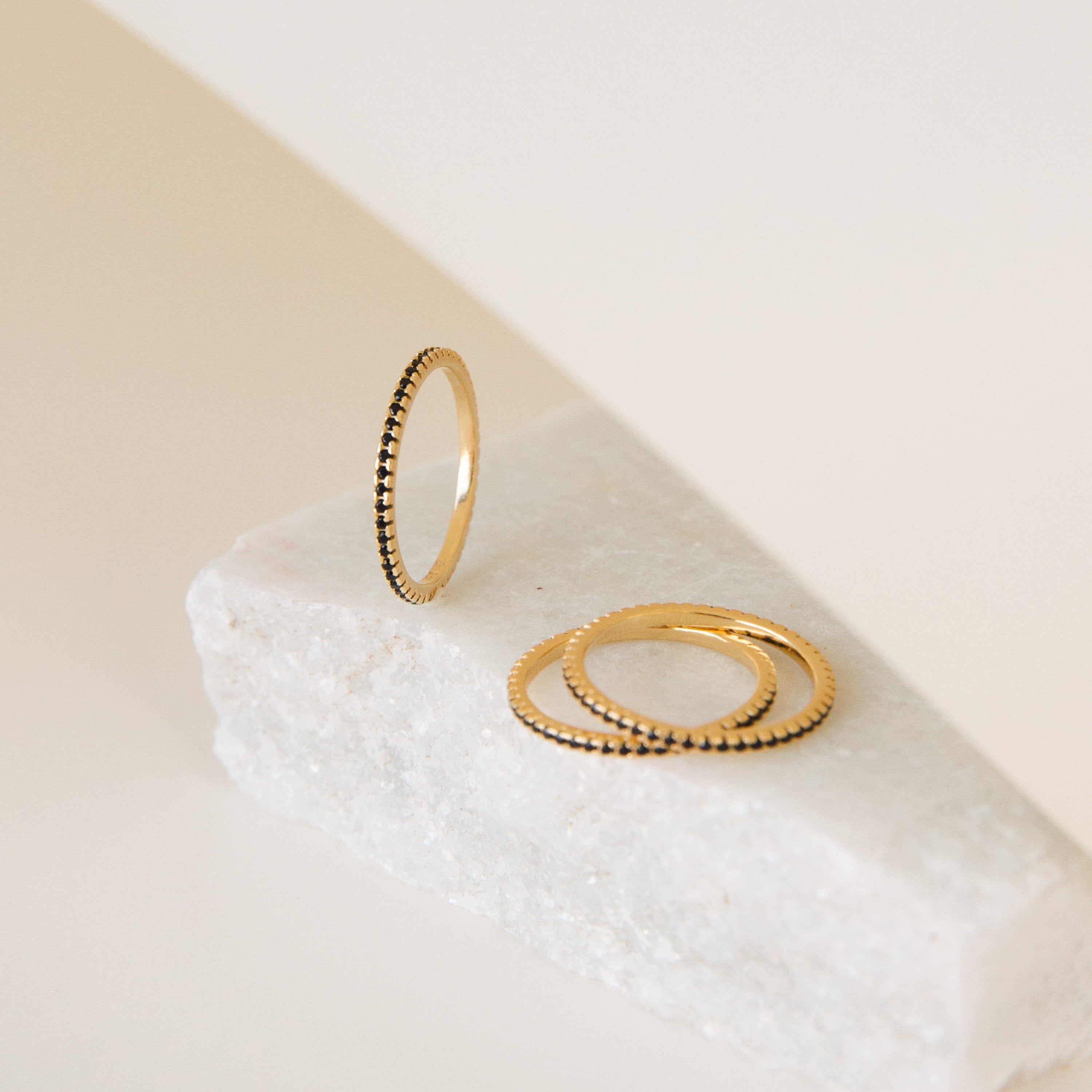 The Amber Eternity Ring, a delicate gold stackable ring with beaded edges, is displayed on a white stone surface against a neutral background.