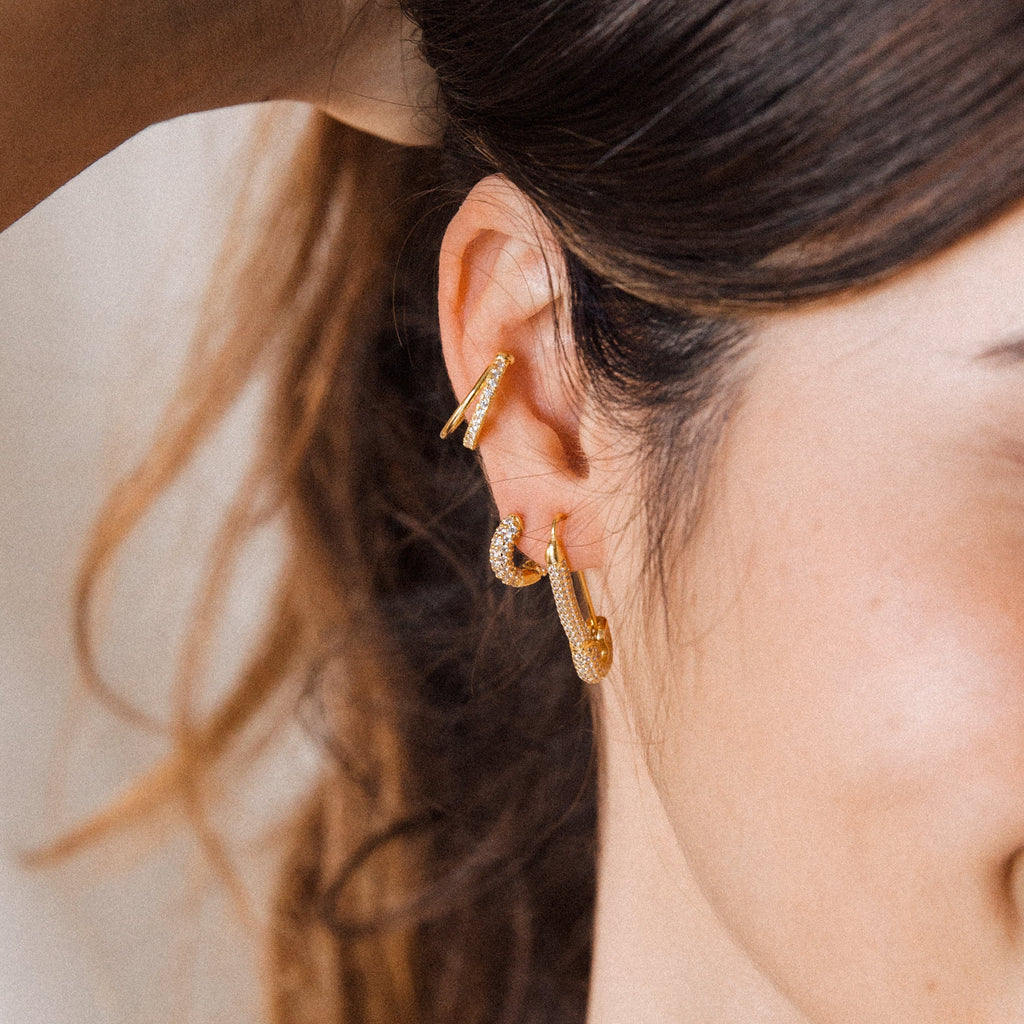A close-up shows a woman's ear with three gold hoops, featuring Kira Pave Huggies, her brown hair tied back and hand resting near her head.