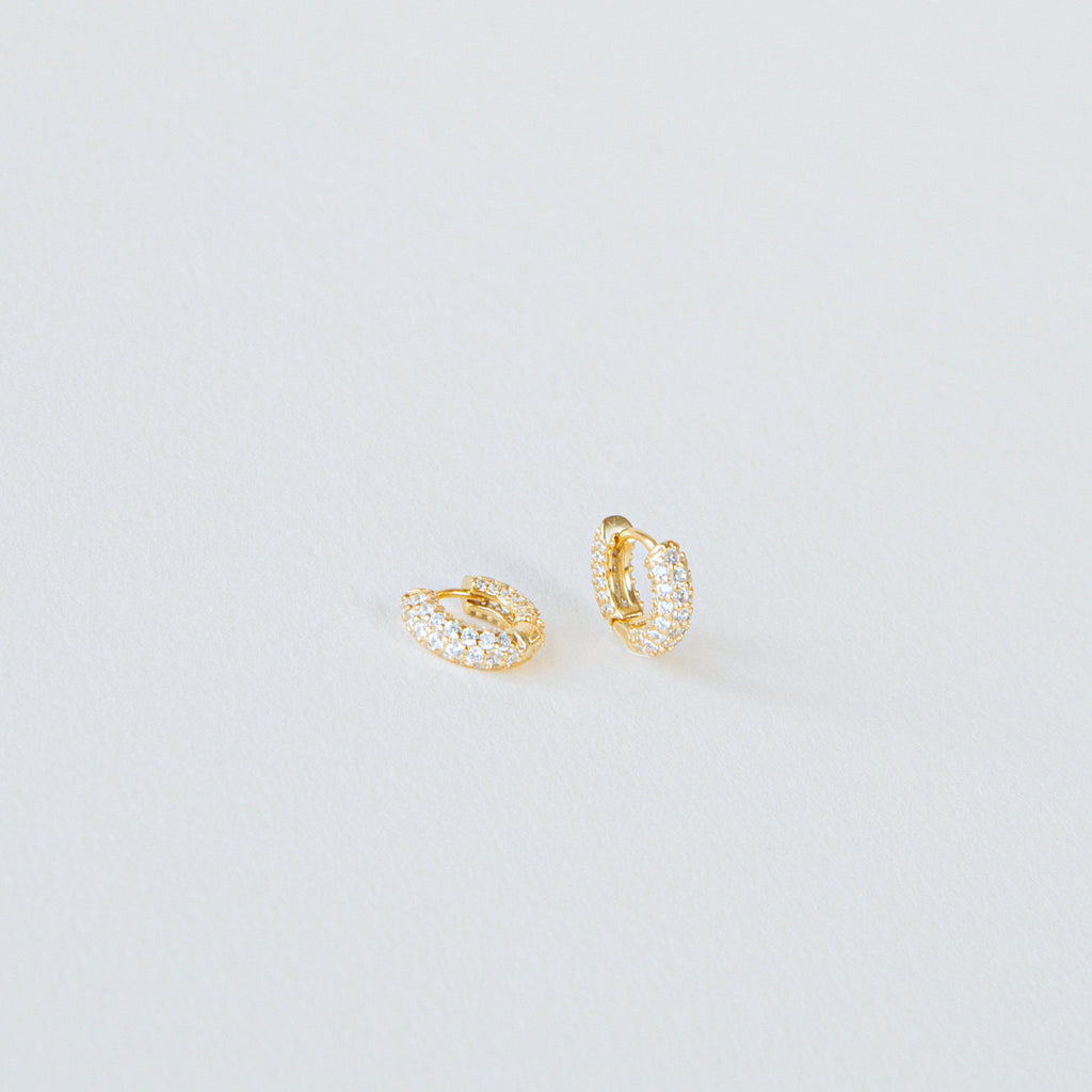 Kira Pave Huggies in small gold feature pavé white stones and are displayed on a plain white surface.