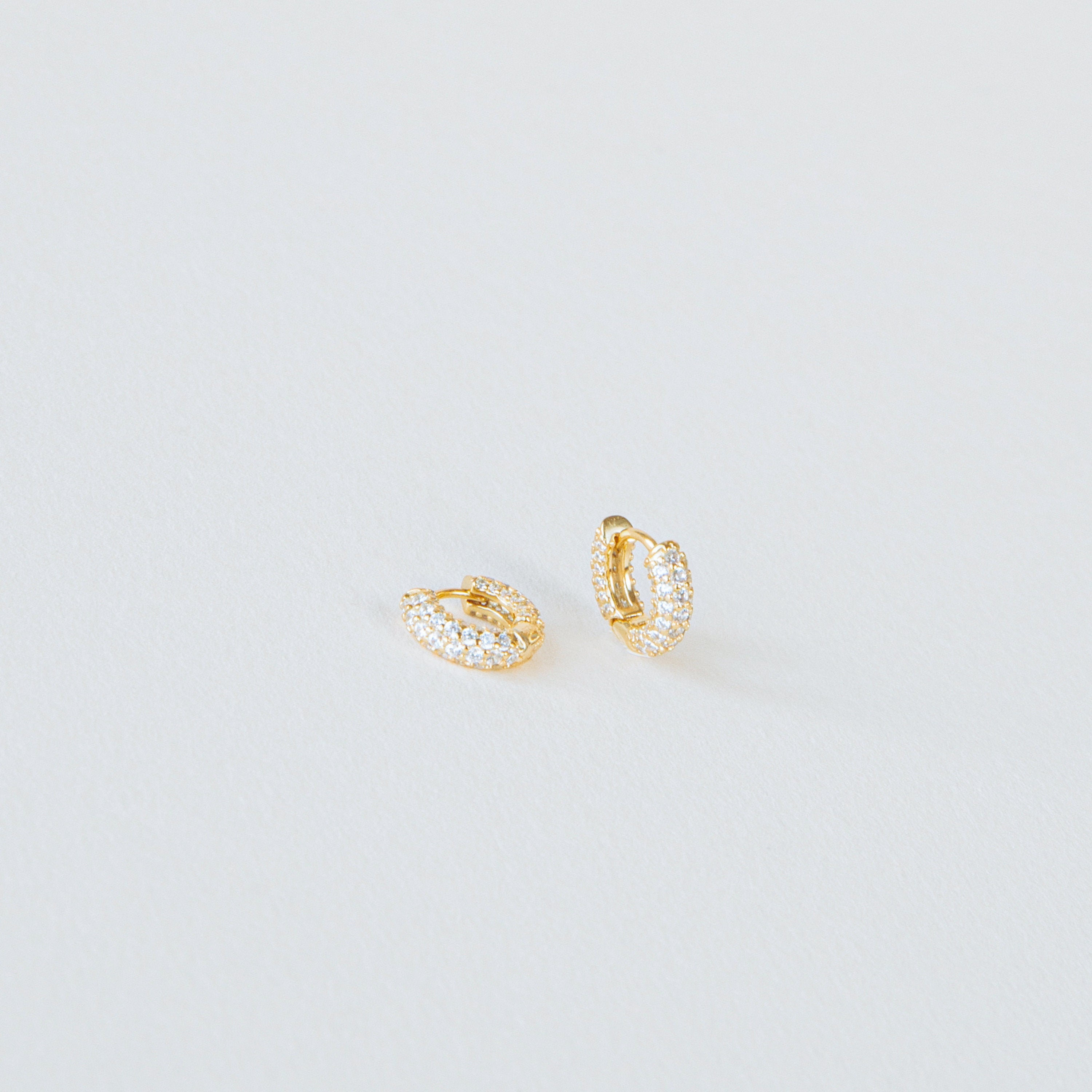 Kira Pave Huggies in small gold feature pavé white stones and are displayed on a plain white surface.