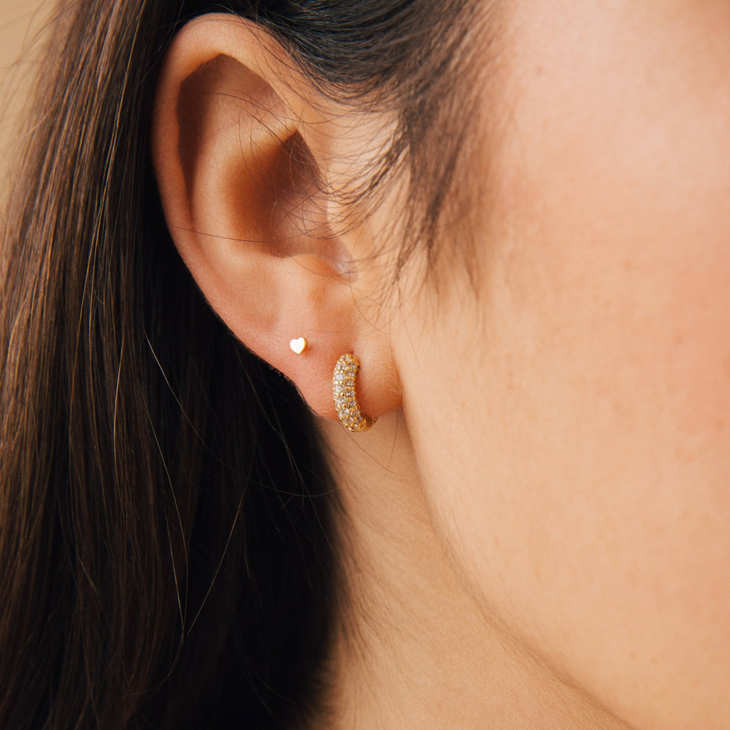 Close-up of a woman's ear wearing Kira Pave Huggies: a pair of shimmering gold hoop earrings adorned with small stones, alongside a small heart-shaped stud.