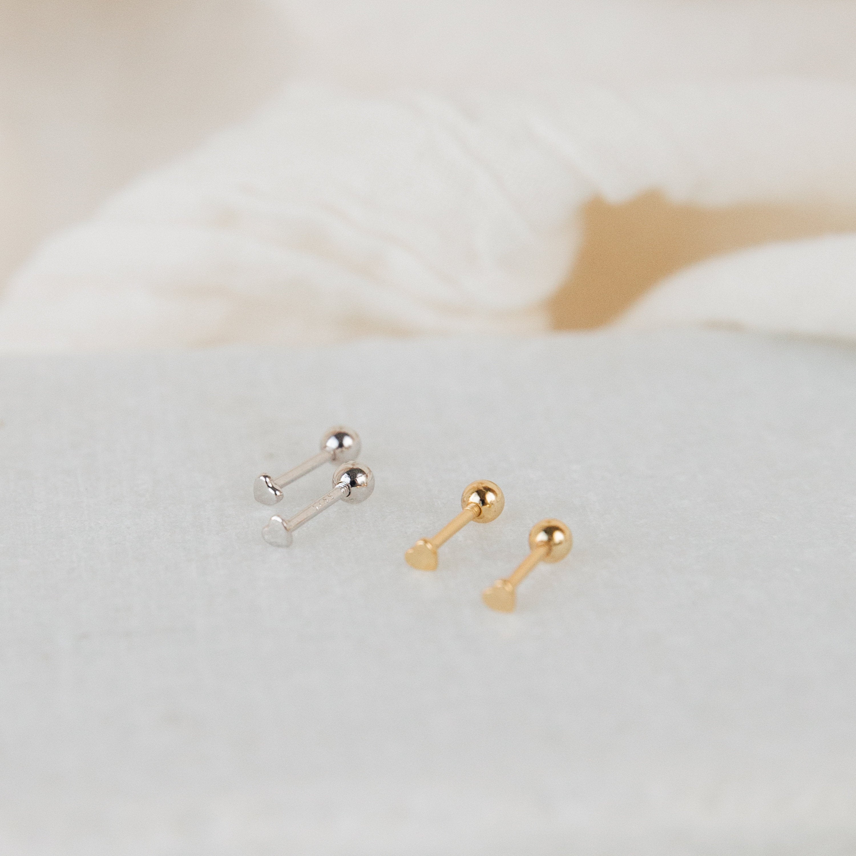 The Tiny Heart Studs in 18K Gold are displayed on a light textured surface with soft cream fabric in the background—making them a chic, minimalist gift choice.