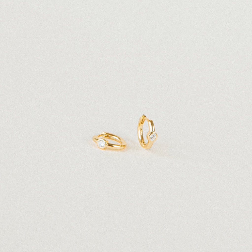 The Aela Huggies are two small gold hoop earrings, each featuring a single clear stone, elegantly displayed on a plain light background.