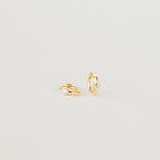 The Aela Huggies are two small gold hoop earrings, each featuring a single clear stone, elegantly displayed on a plain light background.