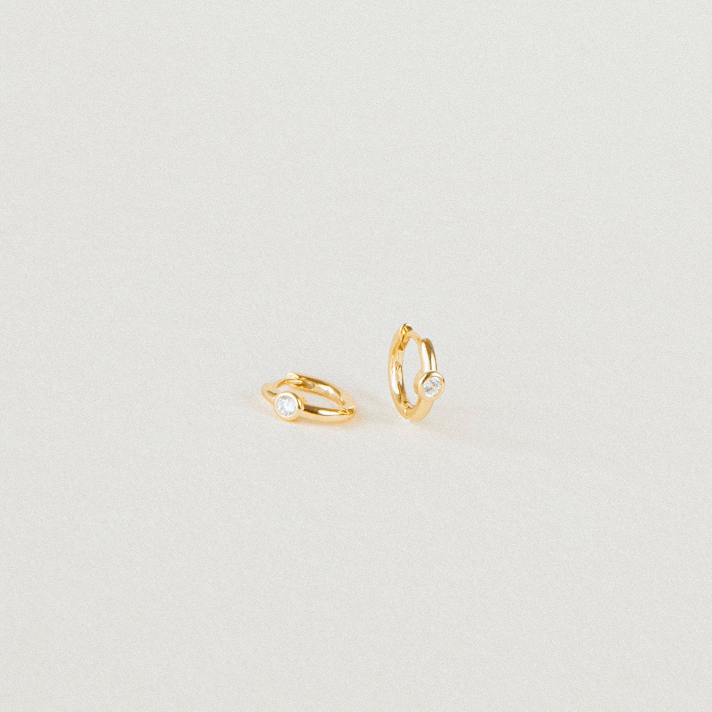 The Aela Huggies are two small gold hoop earrings, each featuring a single clear stone, elegantly displayed on a plain light background.
