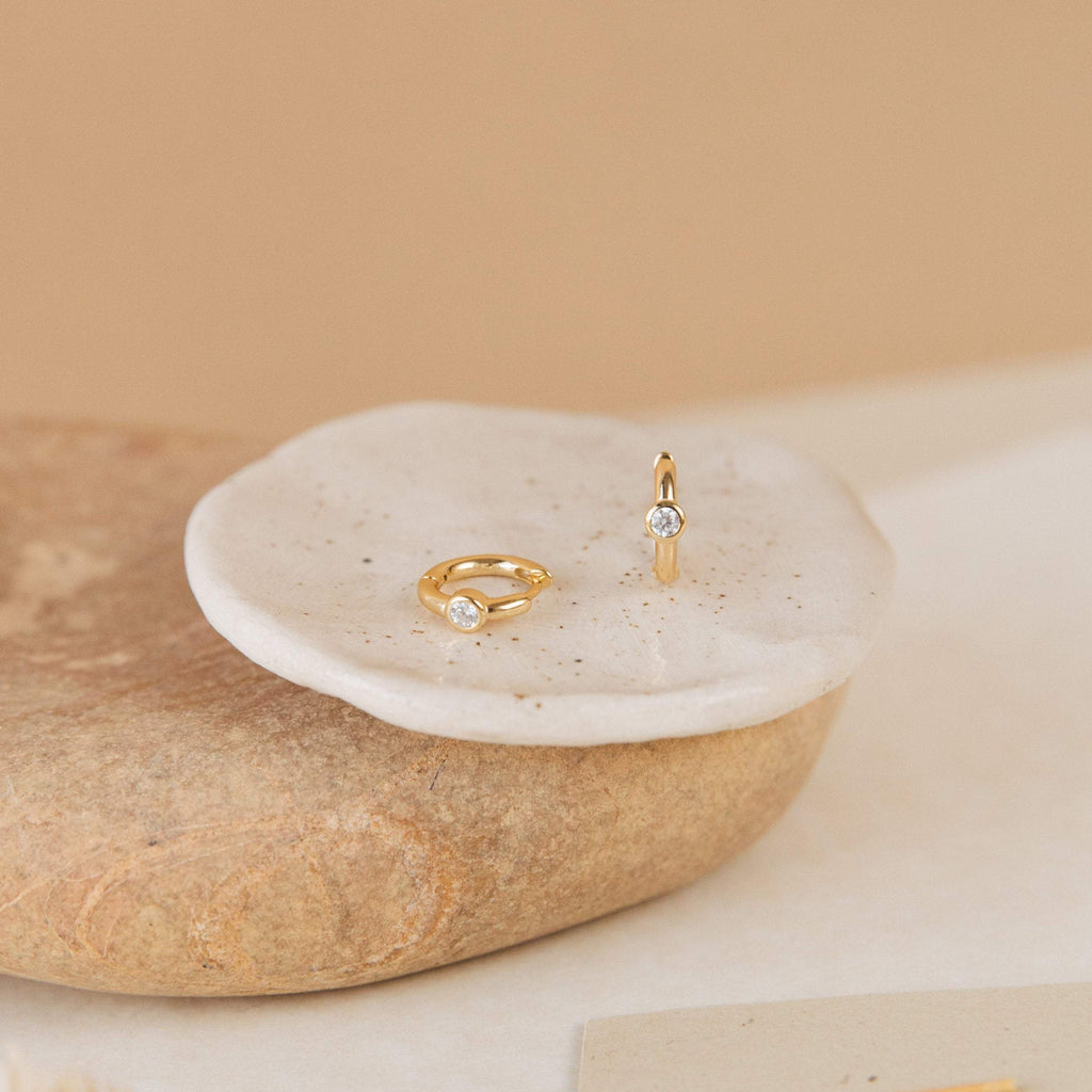 Two gold rings with small clear gemstones rest on a round white dish atop stone, ideal for pairing with the Aela Huggies or other elegant earrings.