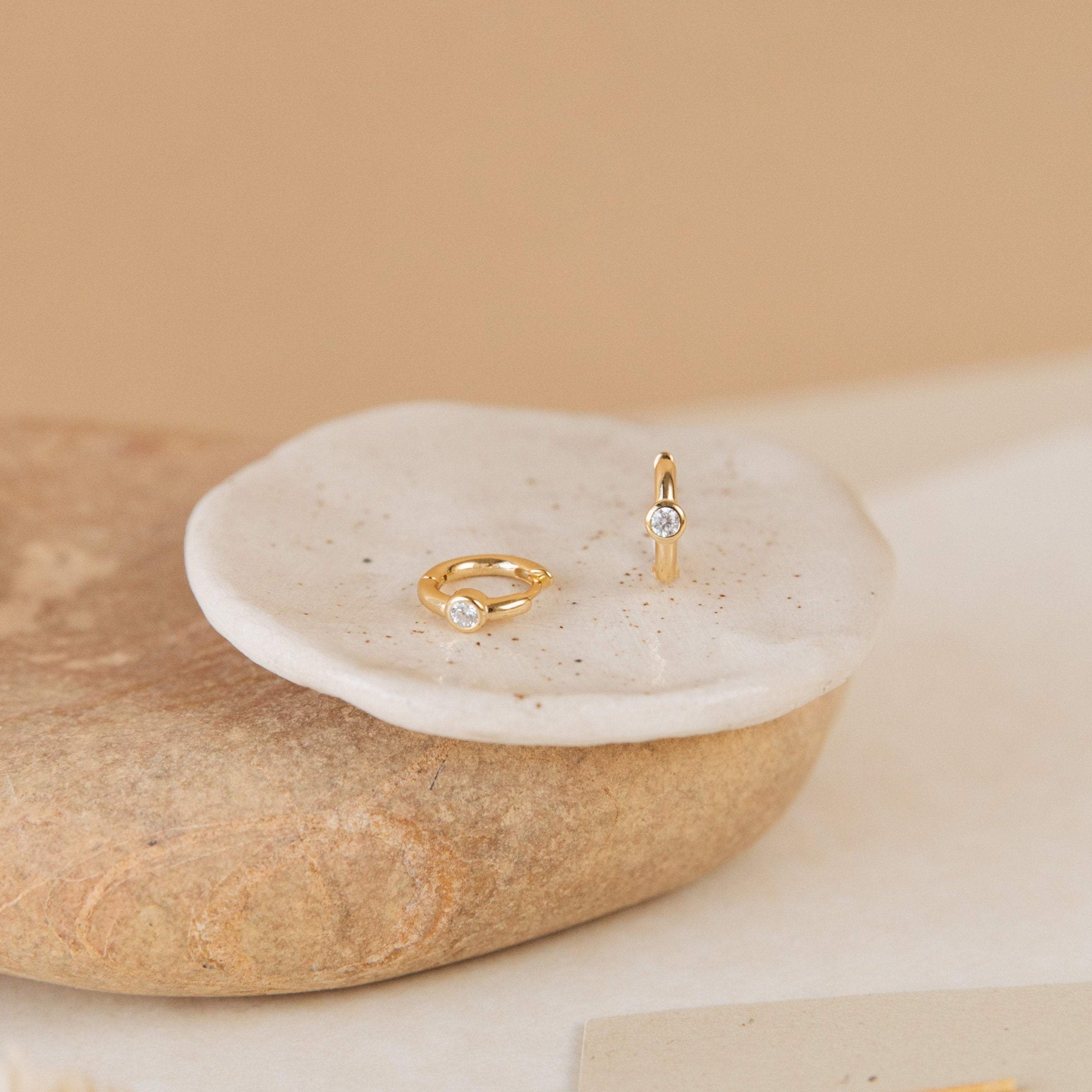 Two gold rings with small clear gemstones rest on a round white dish atop stone, ideal for pairing with the Aela Huggies or other elegant earrings.