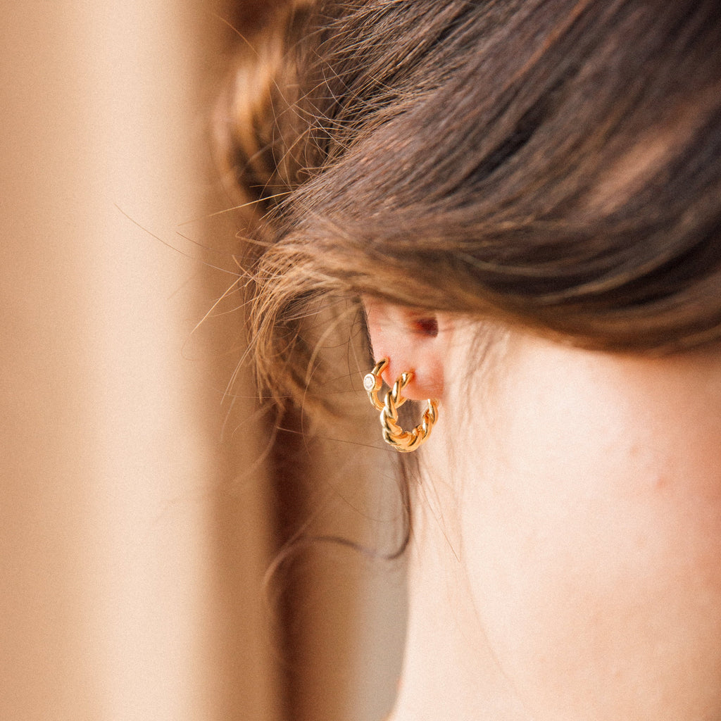 Close-up of a woman’s ear wearing two Aela Huggies gold hoop earrings, styled with loosely pulled back brown hair, highlighting these elegant pieces.