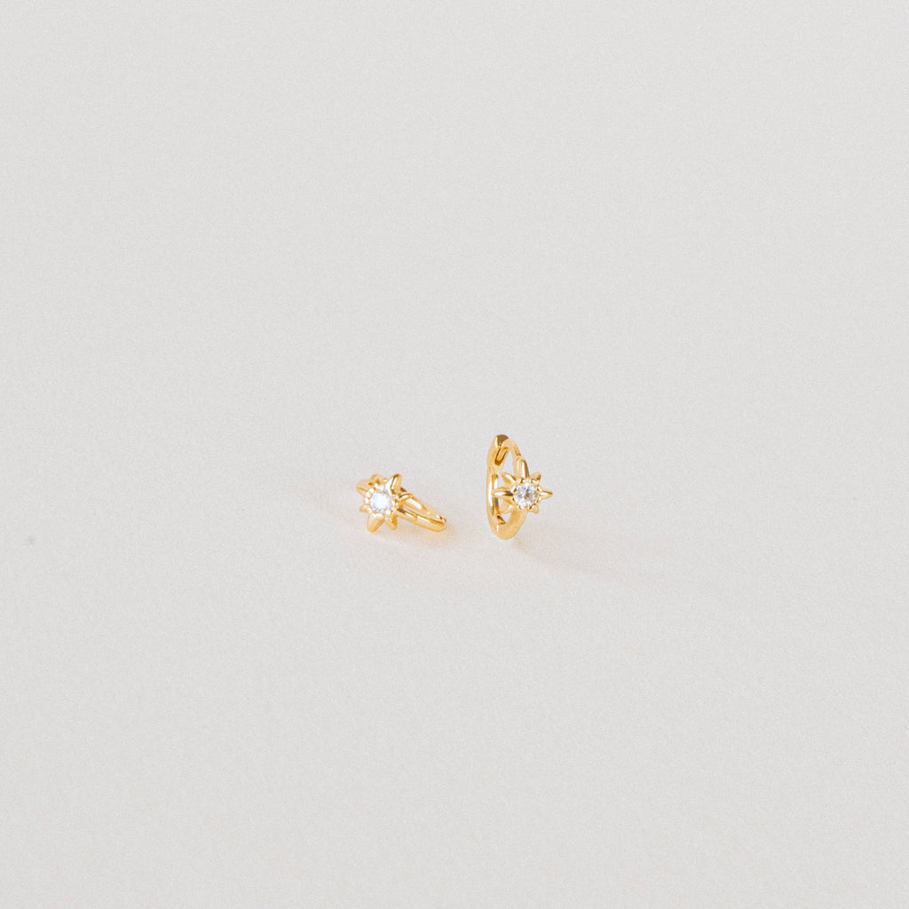 The Enya Huggies are small gold earrings, each with a star-shaped prong design and an iridescent diamond at the center.