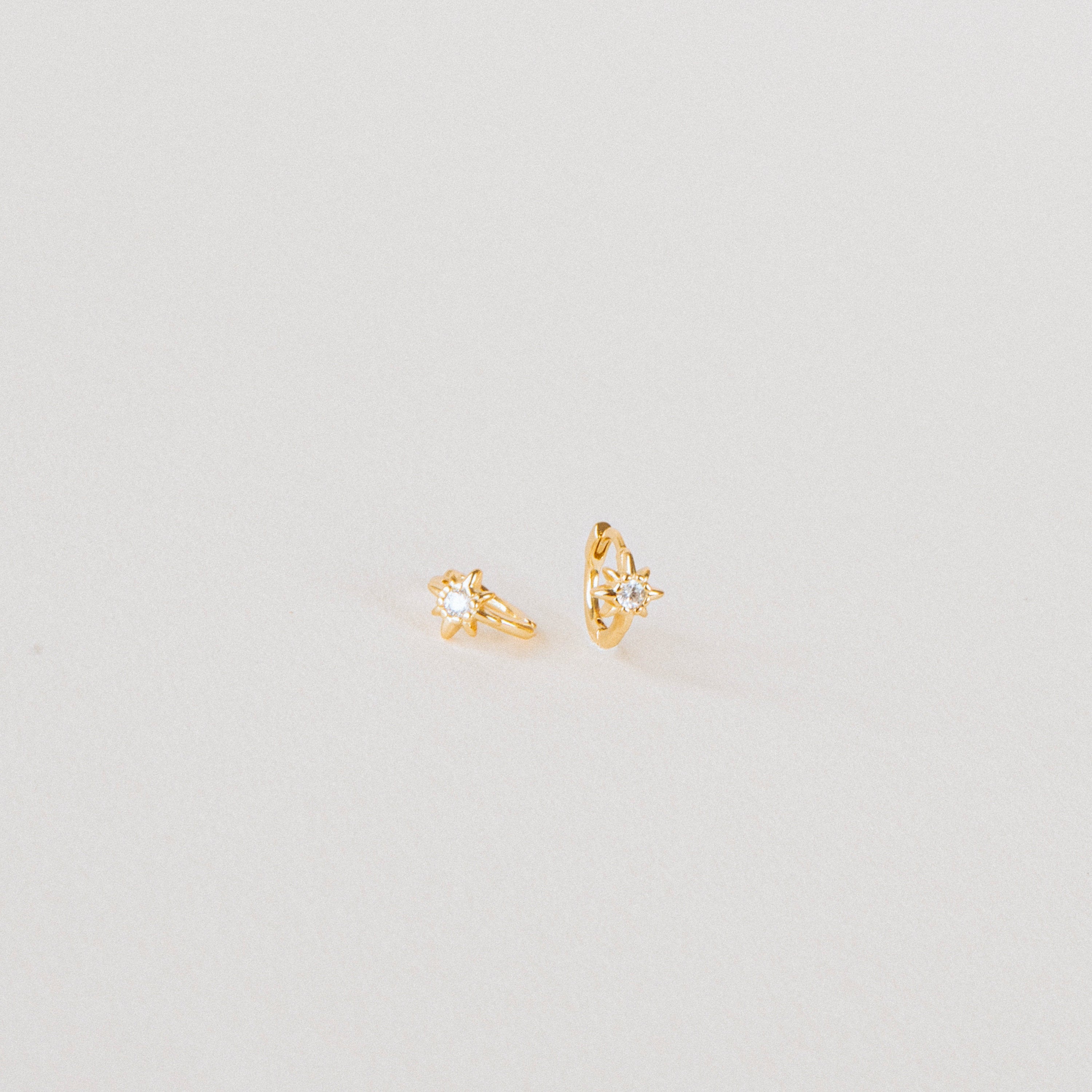 The Enya Huggies are small gold earrings, each with a star-shaped prong design and an iridescent diamond at the center.