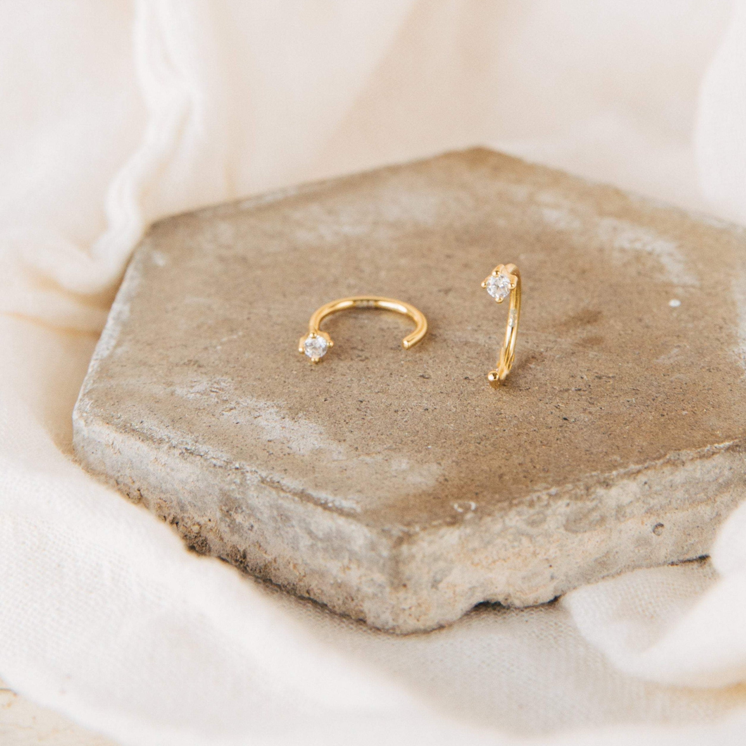 The Saylor Huggies, gold nose rings with small diamonds and dainty hoop earrings, are displayed on a hexagonal stone tile against a cream fabric background.