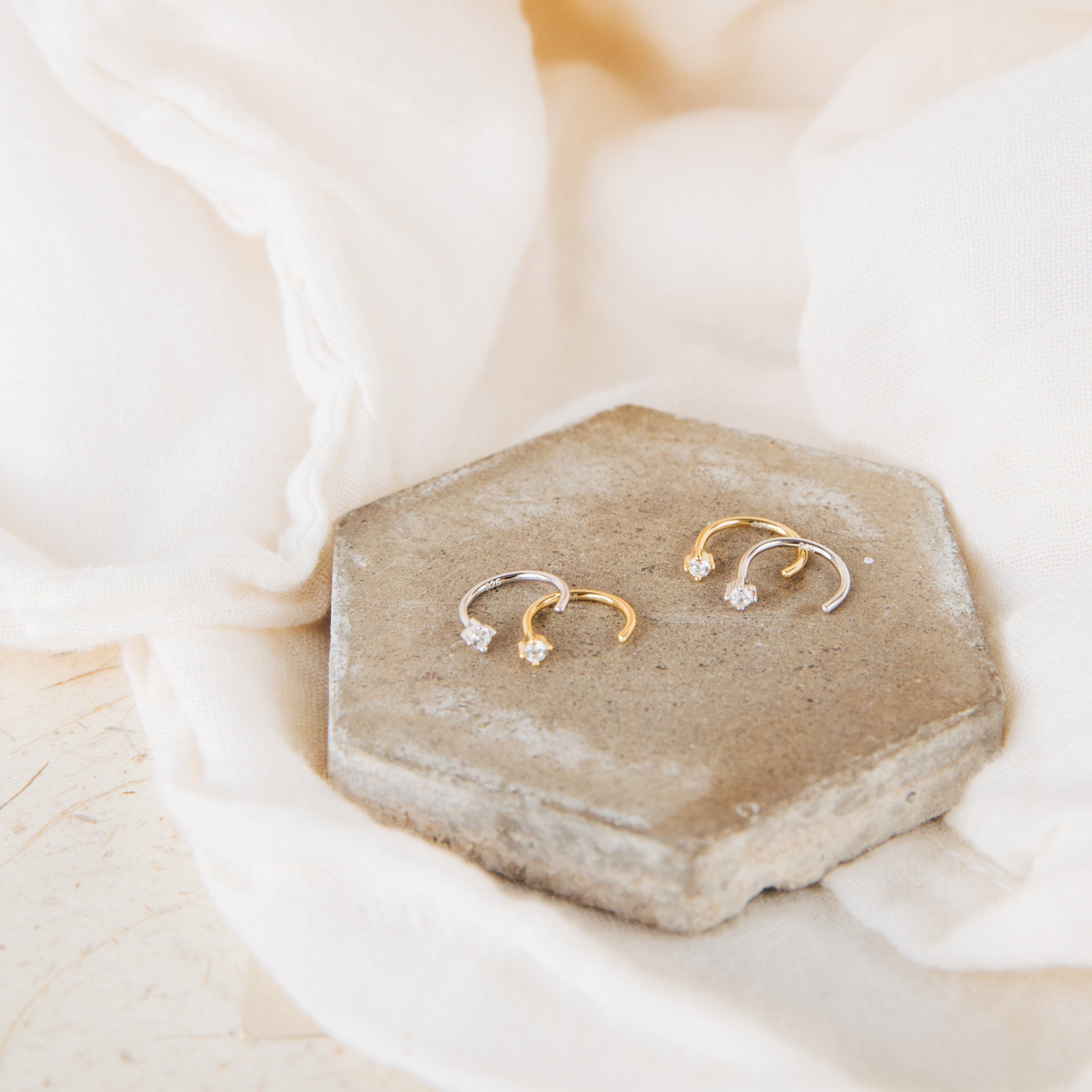 Two pairs of Saylor Huggies—petite, gold and silver dainty hoop earrings with gems—rest on a hexagonal stone atop white fabric.
