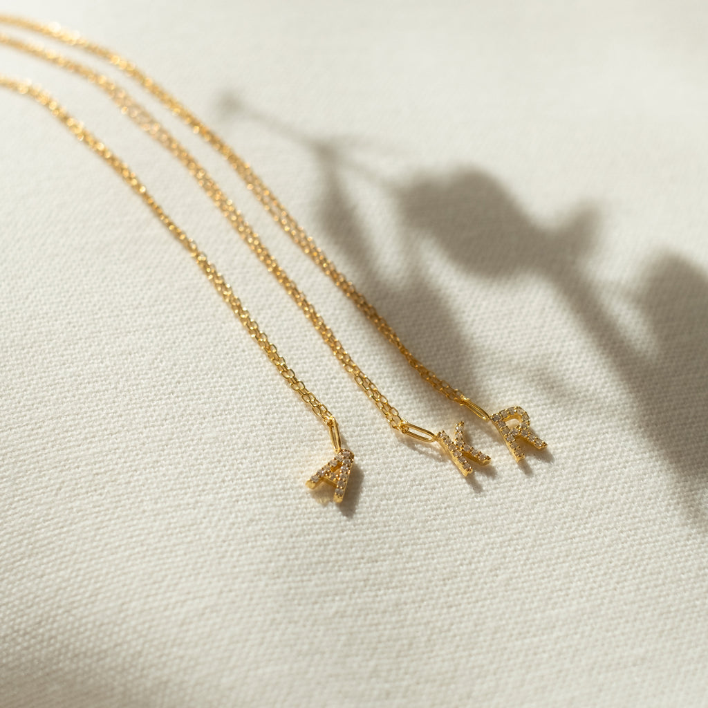 Three gold delicate necklaces, including the Dangling Pave Initial Necklace with a small pendant, are displayed on light textured fabric—an ideal gift for women.