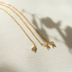 Three gold delicate necklaces, including the Dangling Pave Initial Necklace with a small pendant, are displayed on light textured fabric—an ideal gift for women.
