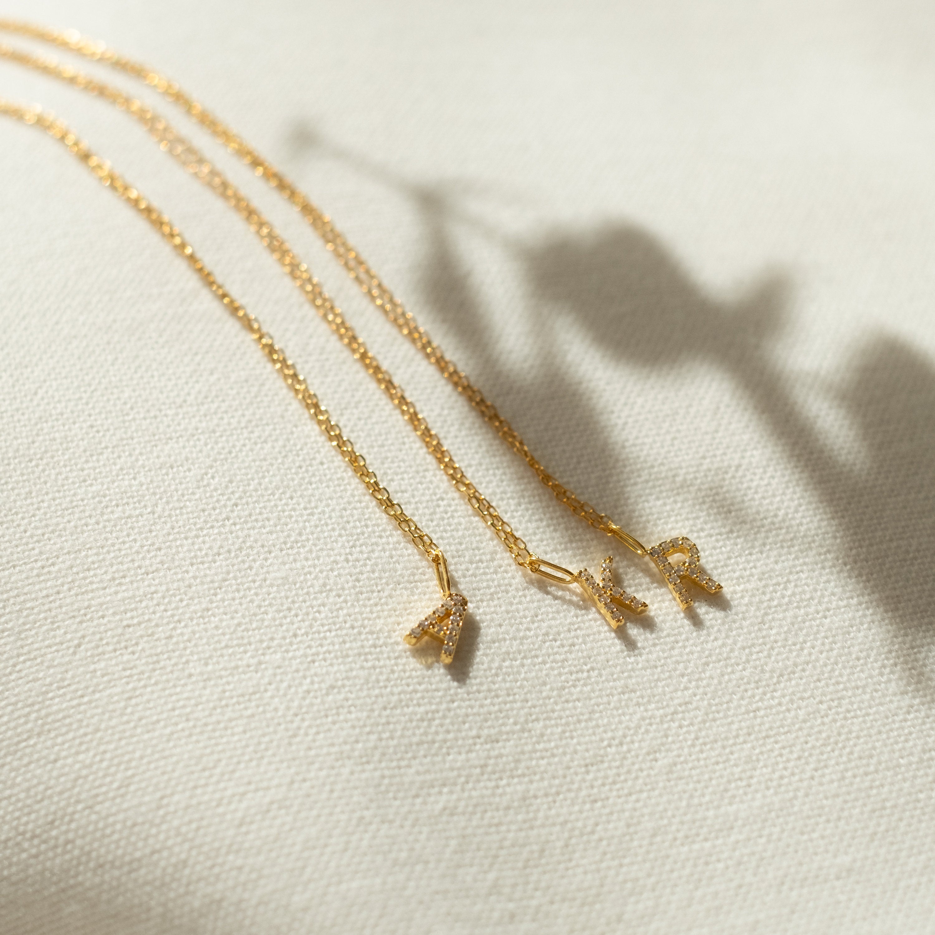 Three gold delicate necklaces, including the Dangling Pave Initial Necklace with a small pendant, are displayed on light textured fabric—an ideal gift for women.