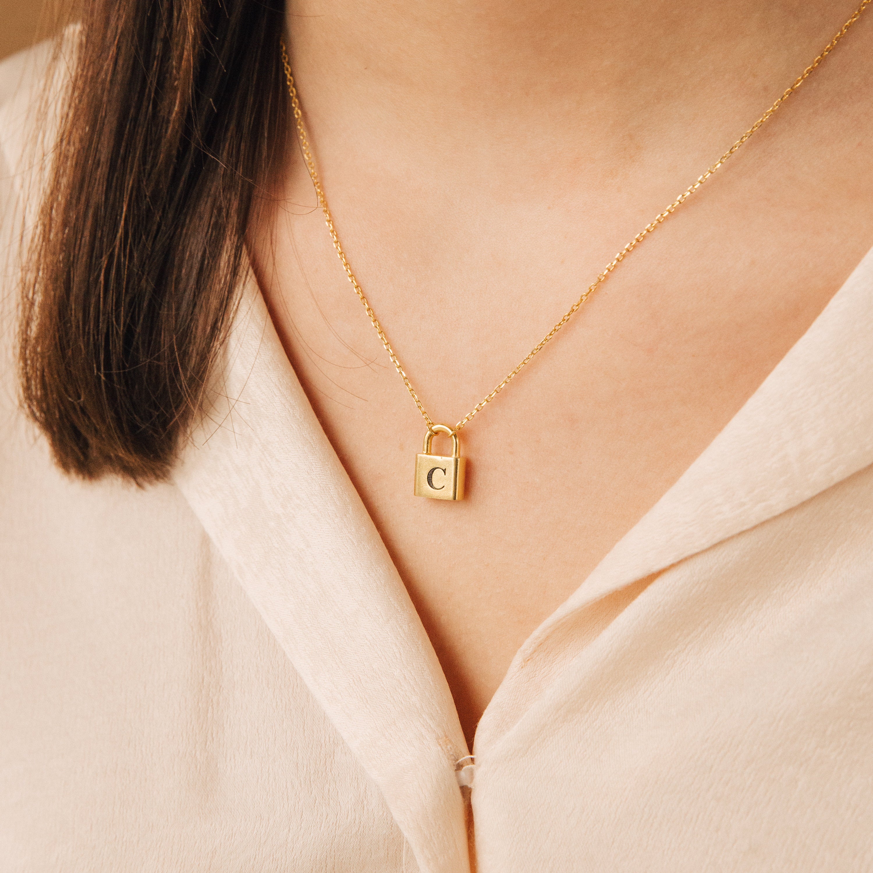 A woman showcases a Padlock Necklace featuring the letter C, adding a stylish accent to her light-colored blouse.