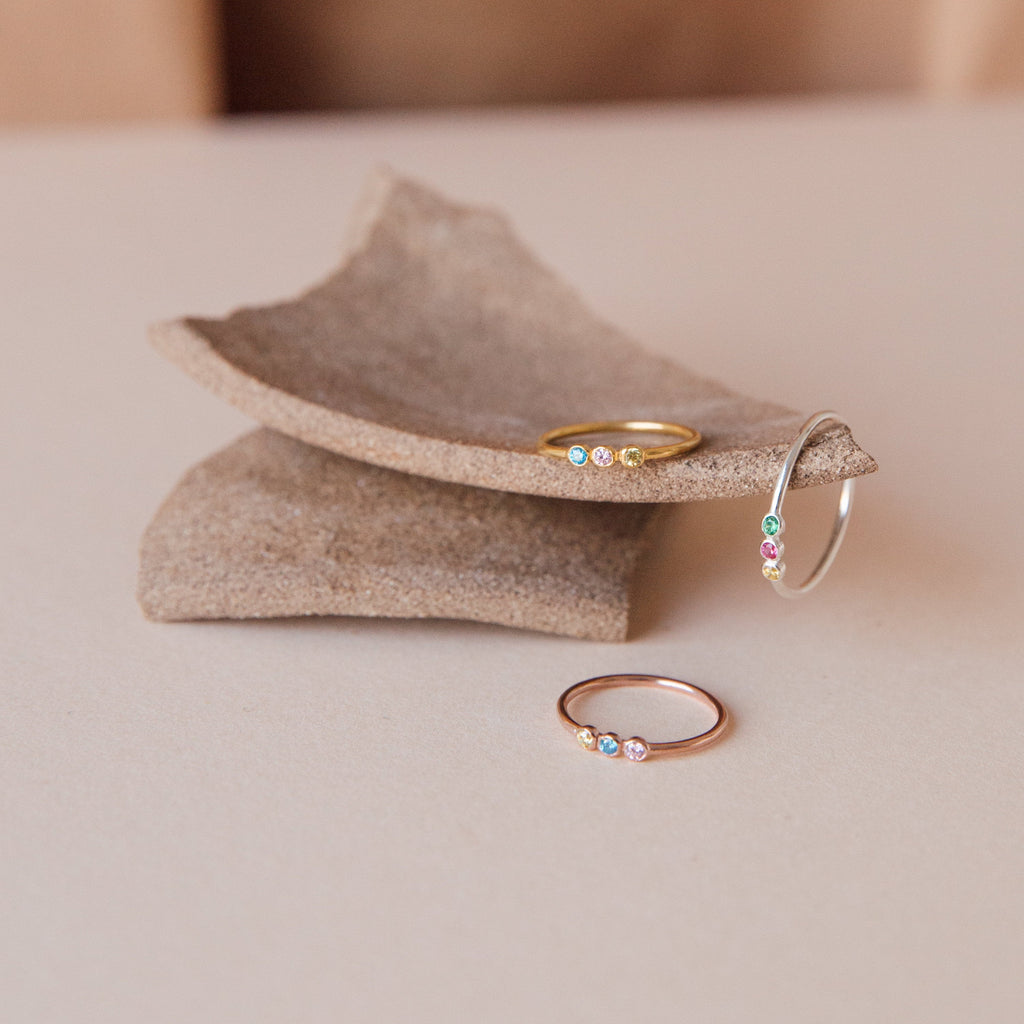 The Tiny Triple Birthstone Ring, a delicate thin band adorned with colorful gemstones, is displayed on textured ceramic pieces.
