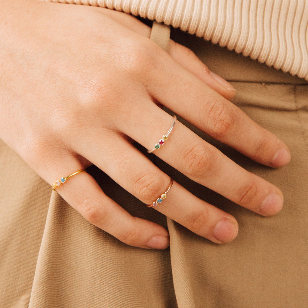 A hand wearing the Tiny Triple Birthstone Ring and two thin bands rests on tan pants and a beige ribbed top.